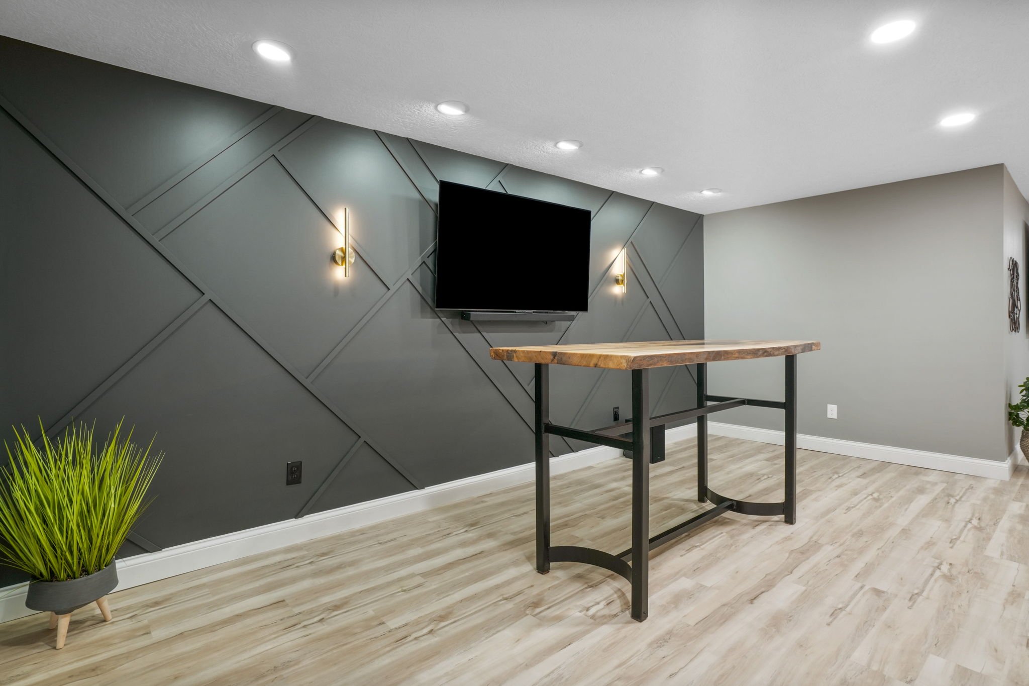 Modern living room with a mounted TV on a geometric gray accent wall, two wall sconces, a wooden table with black metal legs, light wood flooring, a potted green plant, and ceiling recessed lighting.