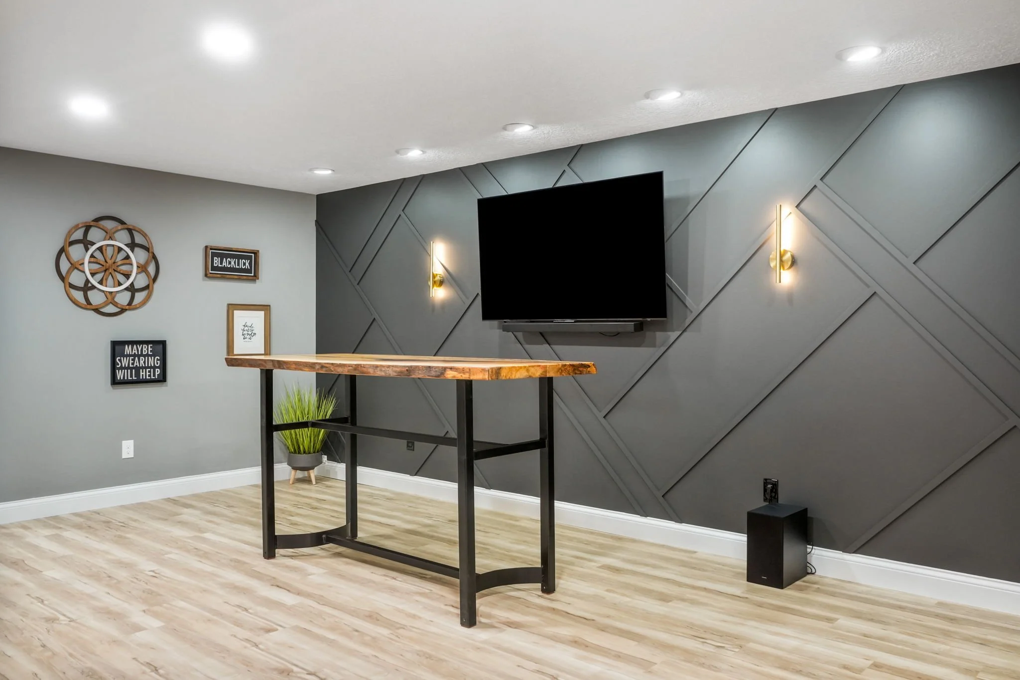 Modern living room with gray geometric accent wall, flat-screen TV, wall sconces, wooden table with black metal frame, potted plant, and framed wall art.