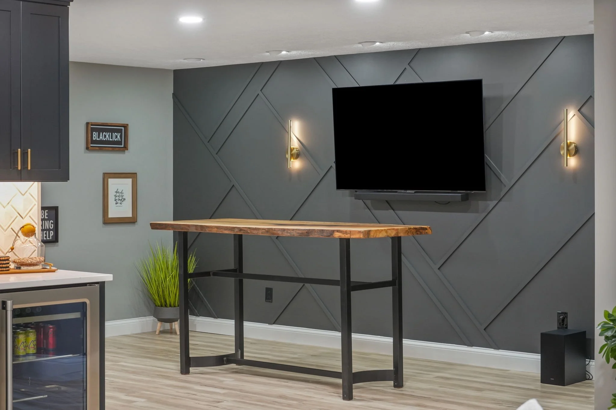 Modern living room with a mounted TV on a gray wall, decorative dark paneling, and two wall sconces. A wooden table with black metal legs is in the center. To the left, a small section of kitchen appliances and counter is visible, with framed wall ar