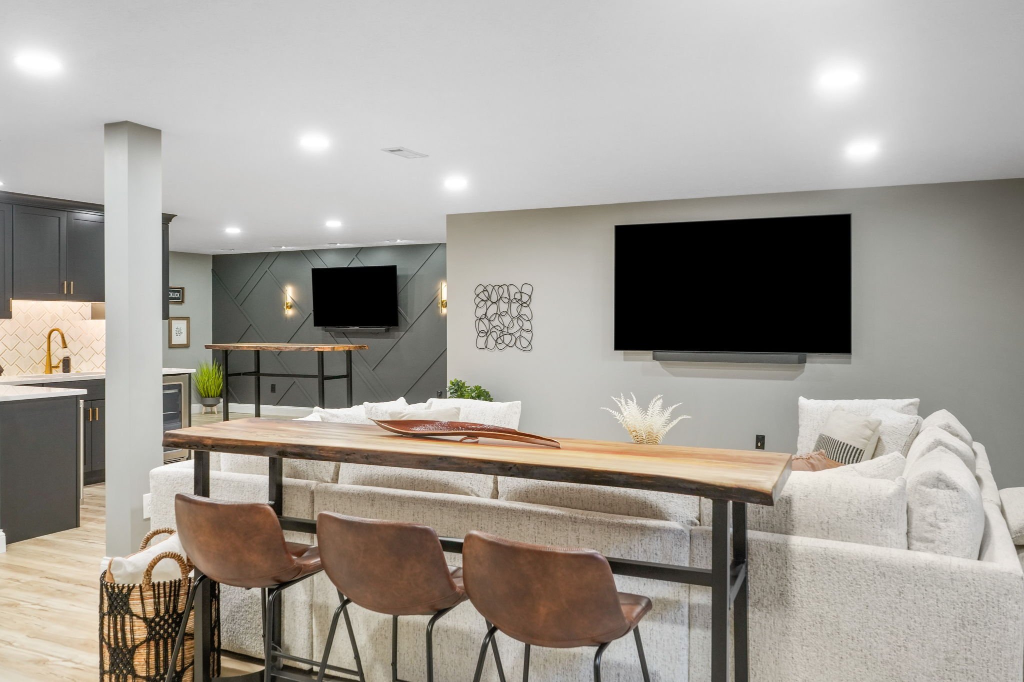 Modern living room with a light-colored sectional sofa, a wooden high-top table, and three brown chairs. Two televisions are mounted on the walls, and a kitchen area with dark cabinetry and a gold faucet is visible in the background. The room feature