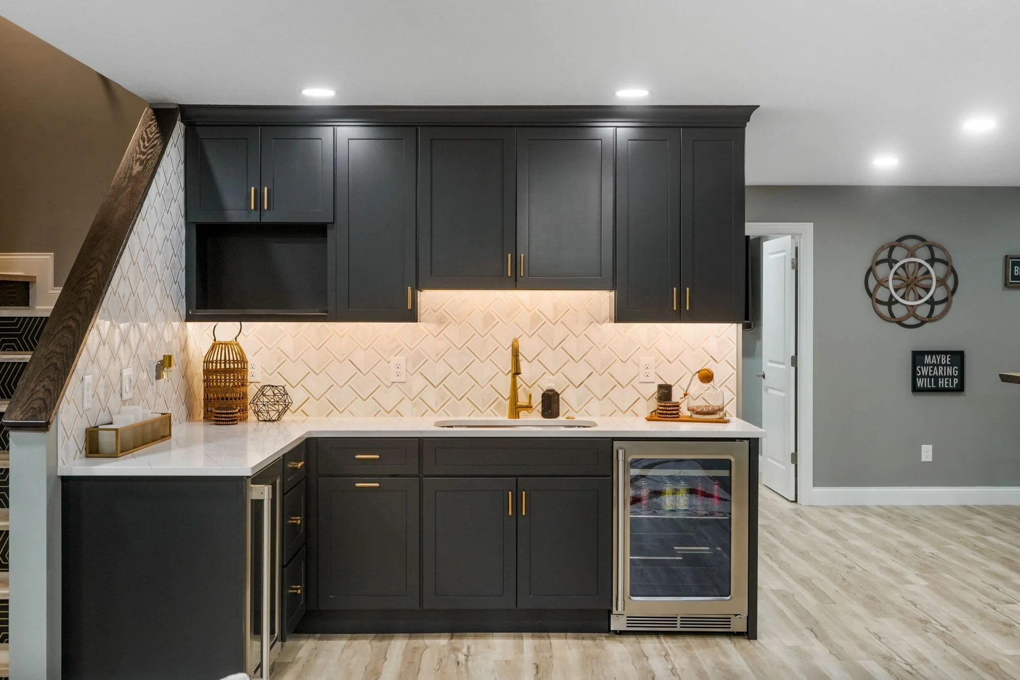 Modern kitchen with dark cabinets, gold handles, white herringbone tile backsplash, white countertop, gold faucet, under-cabinet lighting, small wine fridge, decorative objects, gray wall with framed signs.