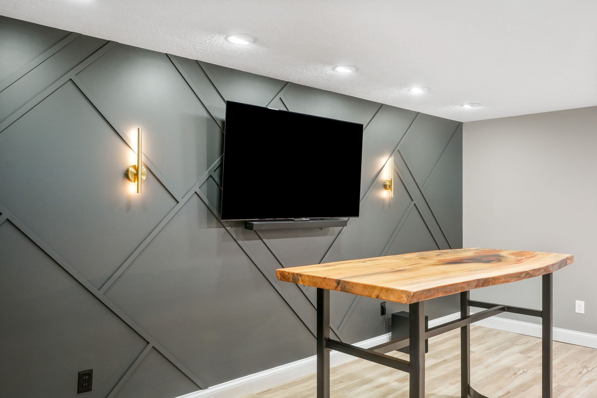 Modern living room corner with a wall-mounted flat-screen TV, gray accented wall with diagonal paneling, two gold wall sconces, and a wooden table with black metal legs.
