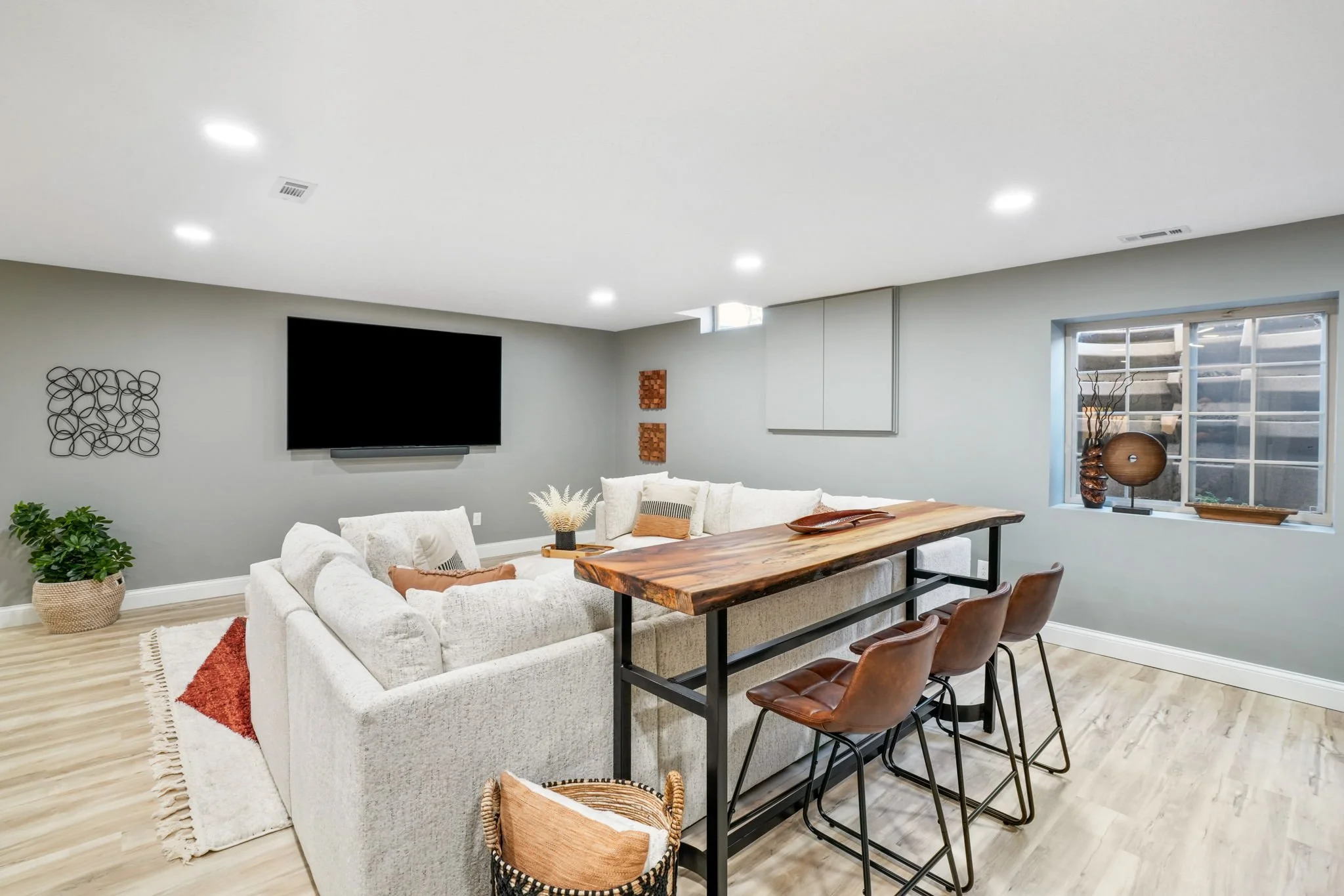 Modern living room with a white sectional sofa, a wooden coffee table, a mounted TV on a gray wall, and a window with decorative vases and plants.