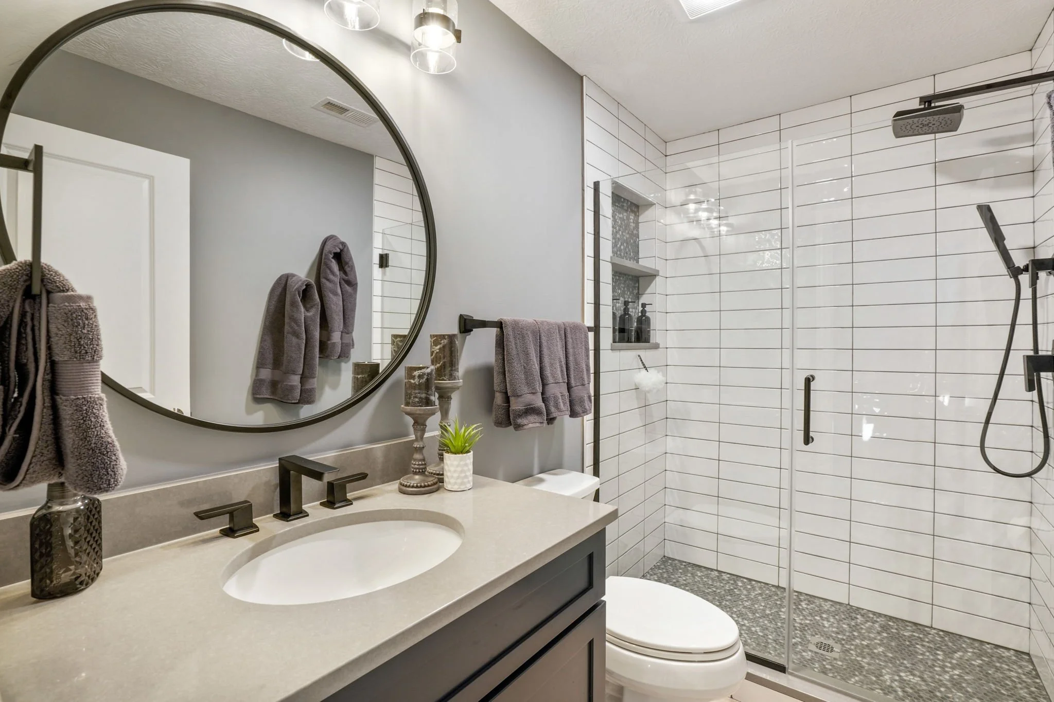 Modern bathroom with a gray vanity, a large oval mirror, gray towels, a small plant, and a walk-in shower with white tile and a rainfall showerhead.