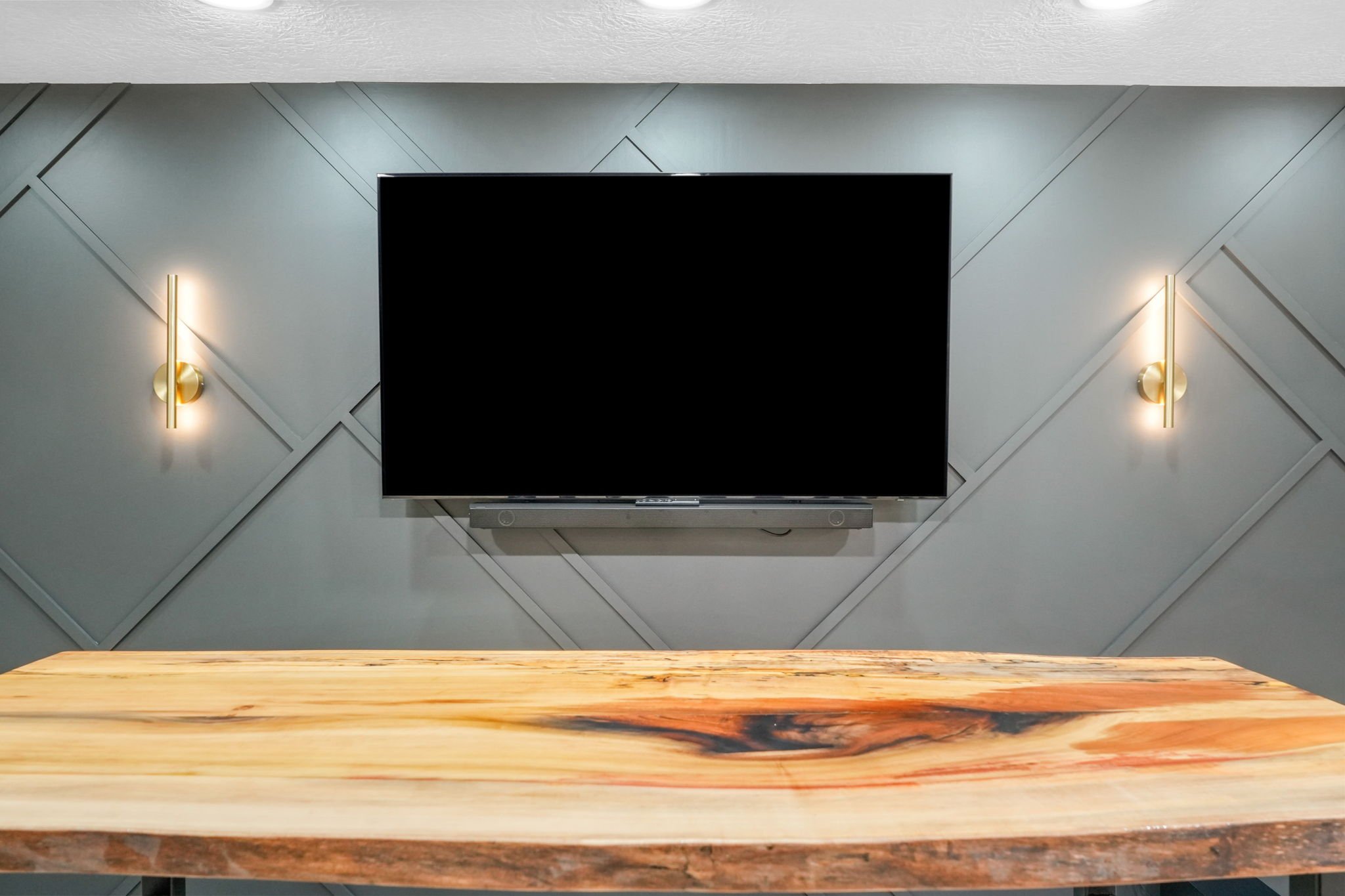 Living room with a mounted flat-screen TV, modern wall sconces, and a wooden table in the foreground.