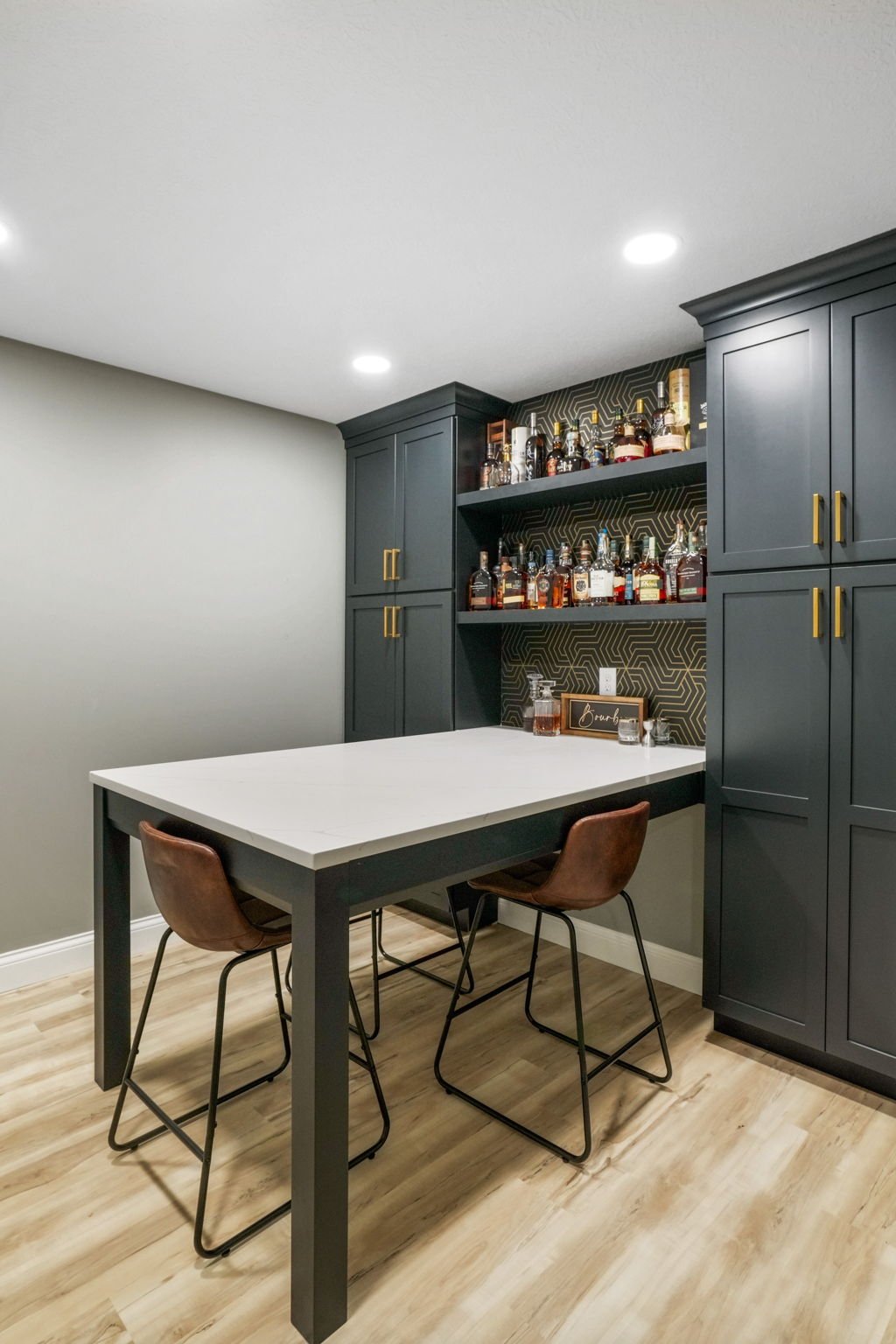 Modern home bar with dark blue cabinetry, open shelves filled with liquor bottles, a white table, and two brown chairs on a wood floor.