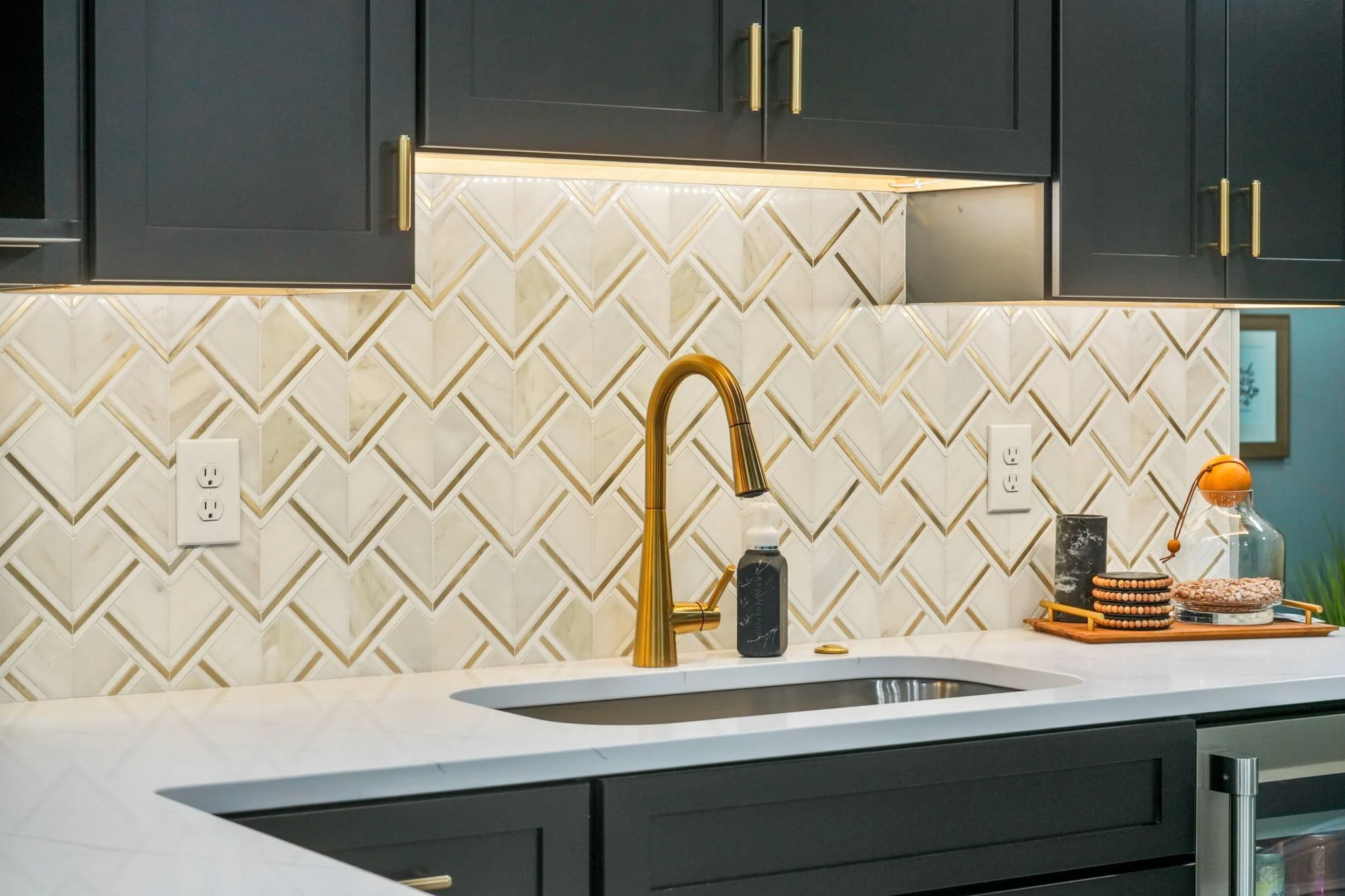 Modern kitchen with white marble countertop, black cabinets with gold handles, and a beige geometric tile backsplash. Gold faucet over sink, and various decorative items on the counter including a black soap dispenser, black plate, and glass jar with