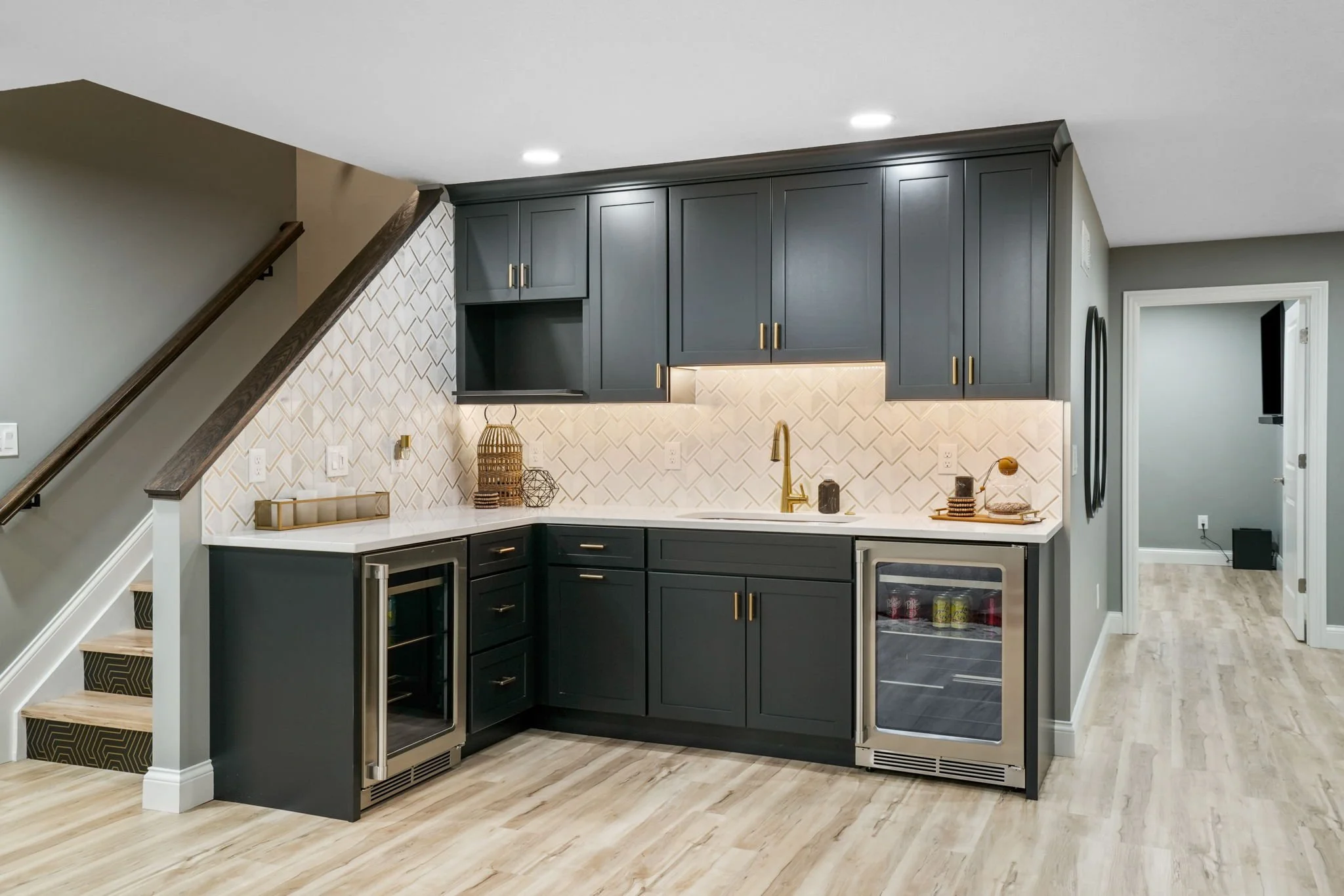 Modern kitchen with black cabinets, white tile backsplash, and built-in mini fridge, under a staircase with light wood flooring.
