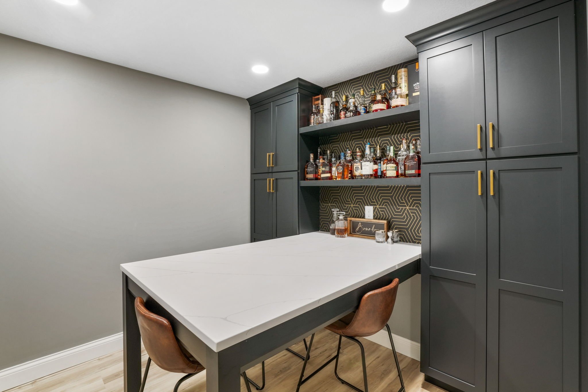 Bar area with dark gray cabinets, white marble countertop, open shelves with bottles of liquor, and a wall with gold and black geometric wallpaper.