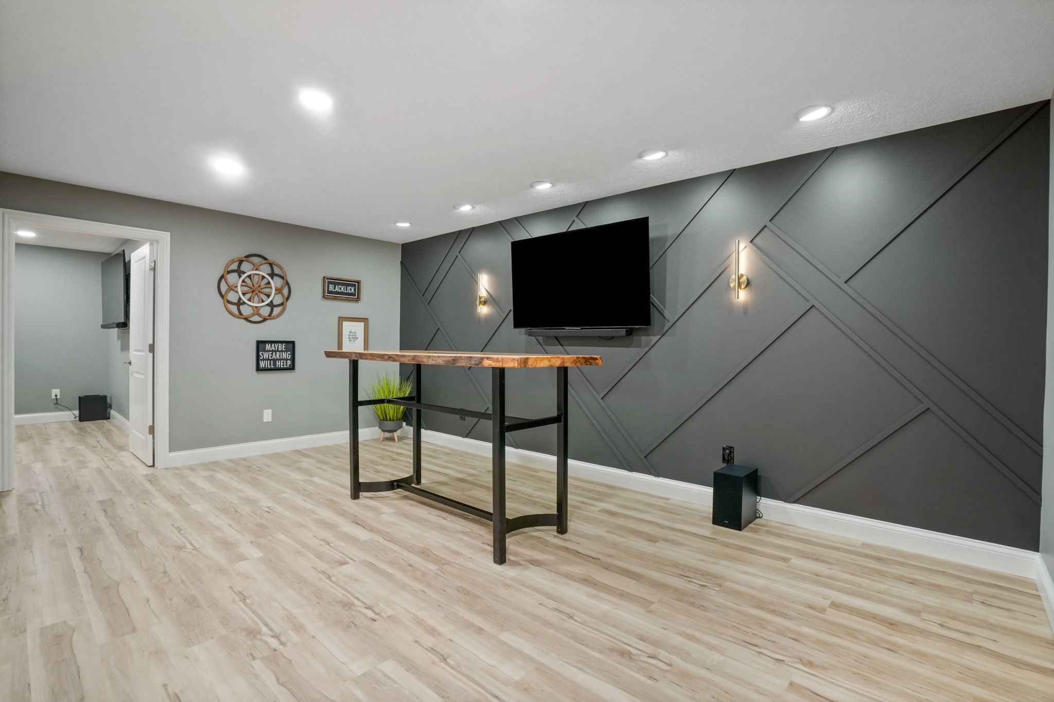 Modern living room with a mounted flat-screen TV on a gray zebra wood accent wall, a tall wooden table, a small green plant, and wall lights, with light wood flooring and a doorway leading to another room.
