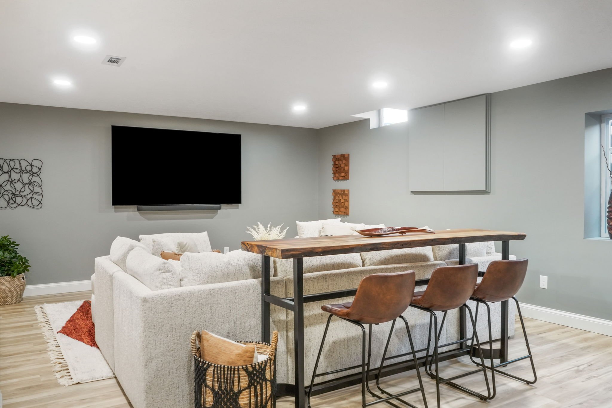 Modern living room with a large white sectional sofa, a wooden bar-height table with three brown chairs, a wall-mounted flat-screen TV, plants, and decorative wall art.