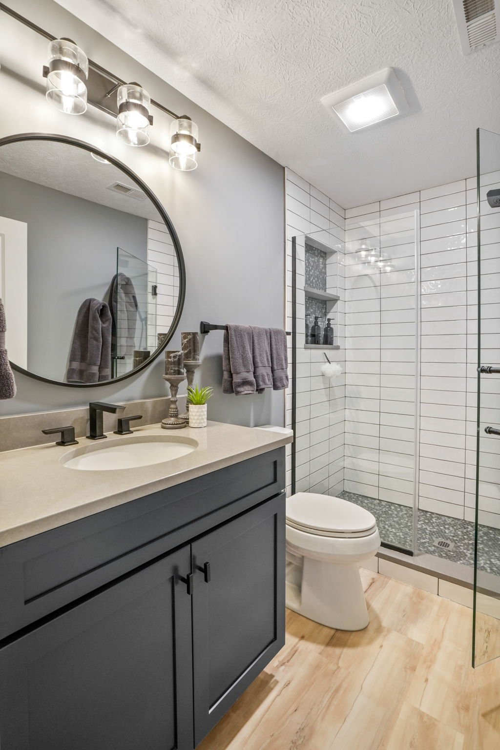 Modern bathroom with gray vanity, beige countertop, round mirror, gray towels, plants, and a walk-in shower with white subway tiles, built-in niche, and pebble floor.