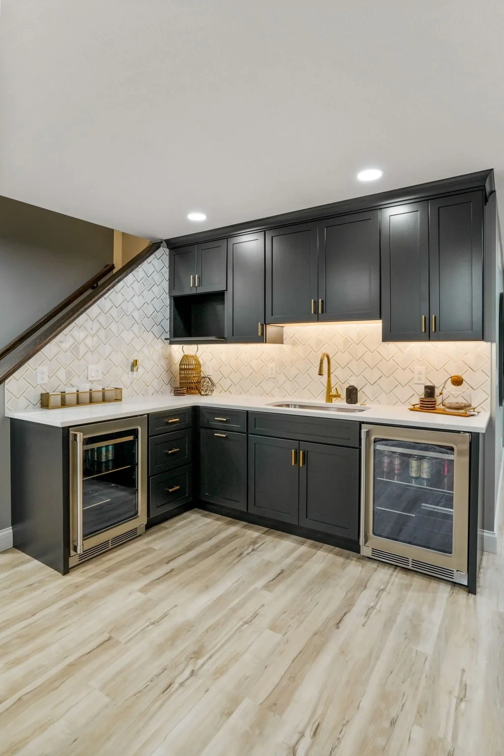 Modern kitchen with black cabinets, white backsplash, and gold hardware, featuring a wine fridge and mini fridge, under- cabinet lighting, and decor accents on the counter.