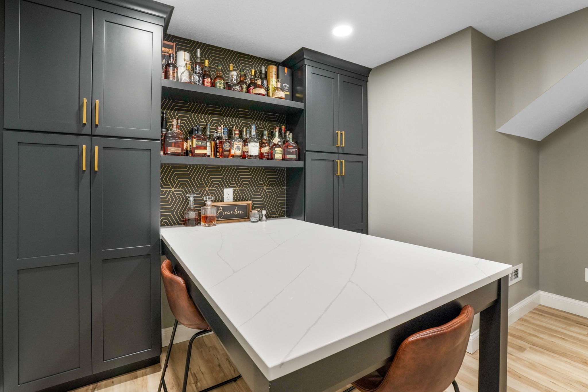 A home bar area with black cabinets, a white marble countertop, and a shelf with various bottles of alcohol, set against a decorative gold and black geometric wallpaper.