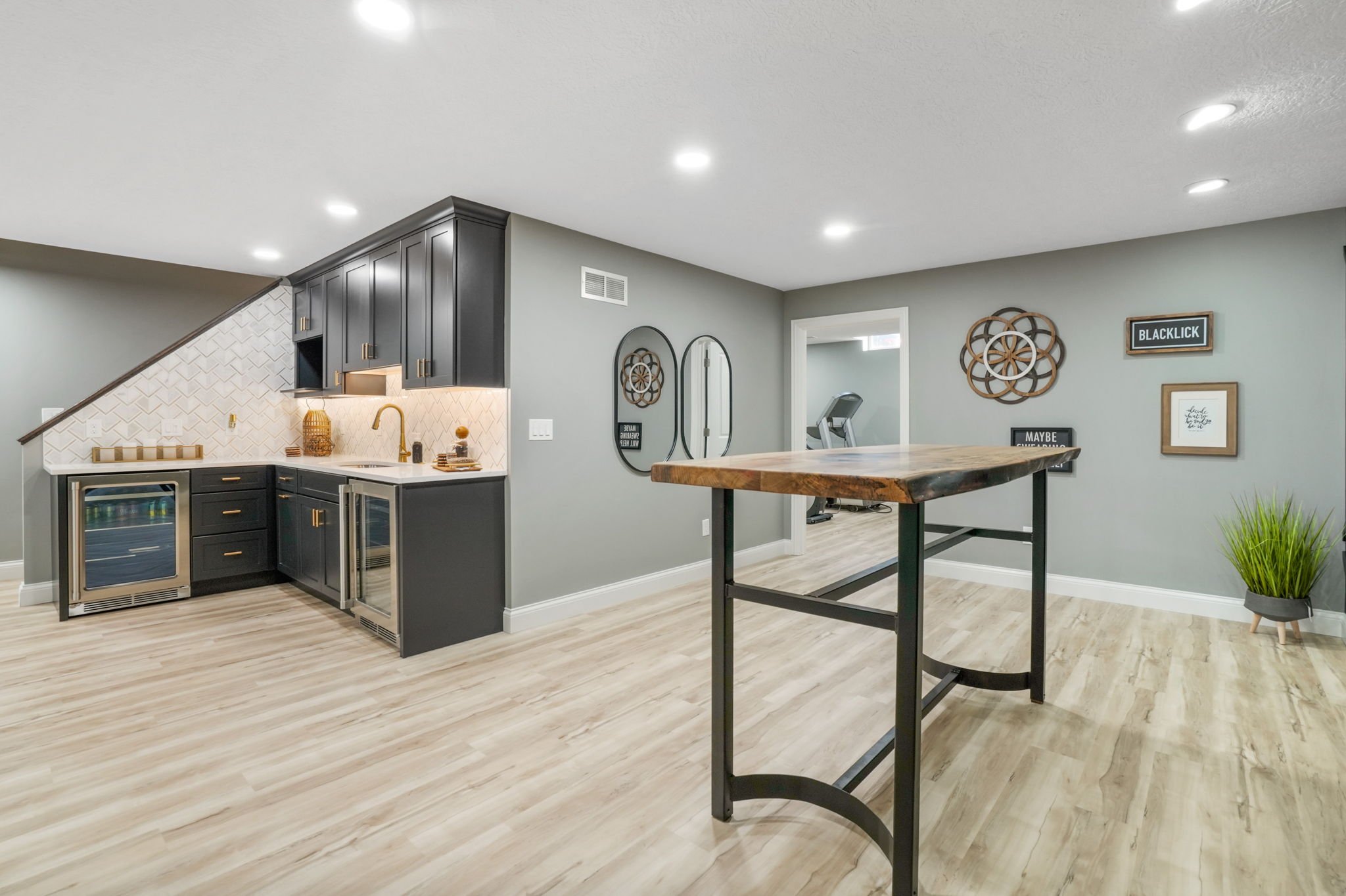 Modern basement kitchen with black cabinets, a small bar area, and wooden accents. Light hardwood floors, green walls, and decorative wall art. Small kitchenette with a gold faucet and white backsplash.