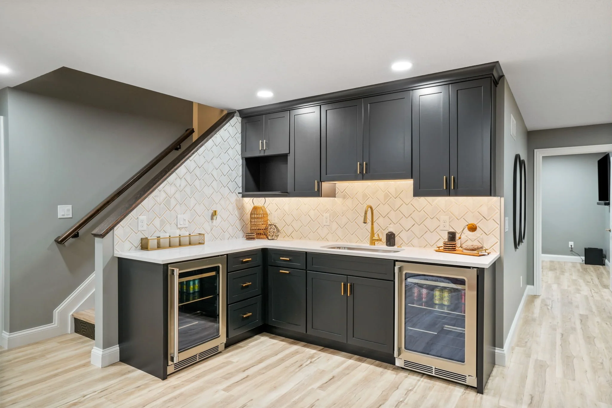 Modern kitchen with dark gray cabinets, white countertop, gold hardware, and a white tile backsplash. Includes small wine fridges and minimal decorative items.