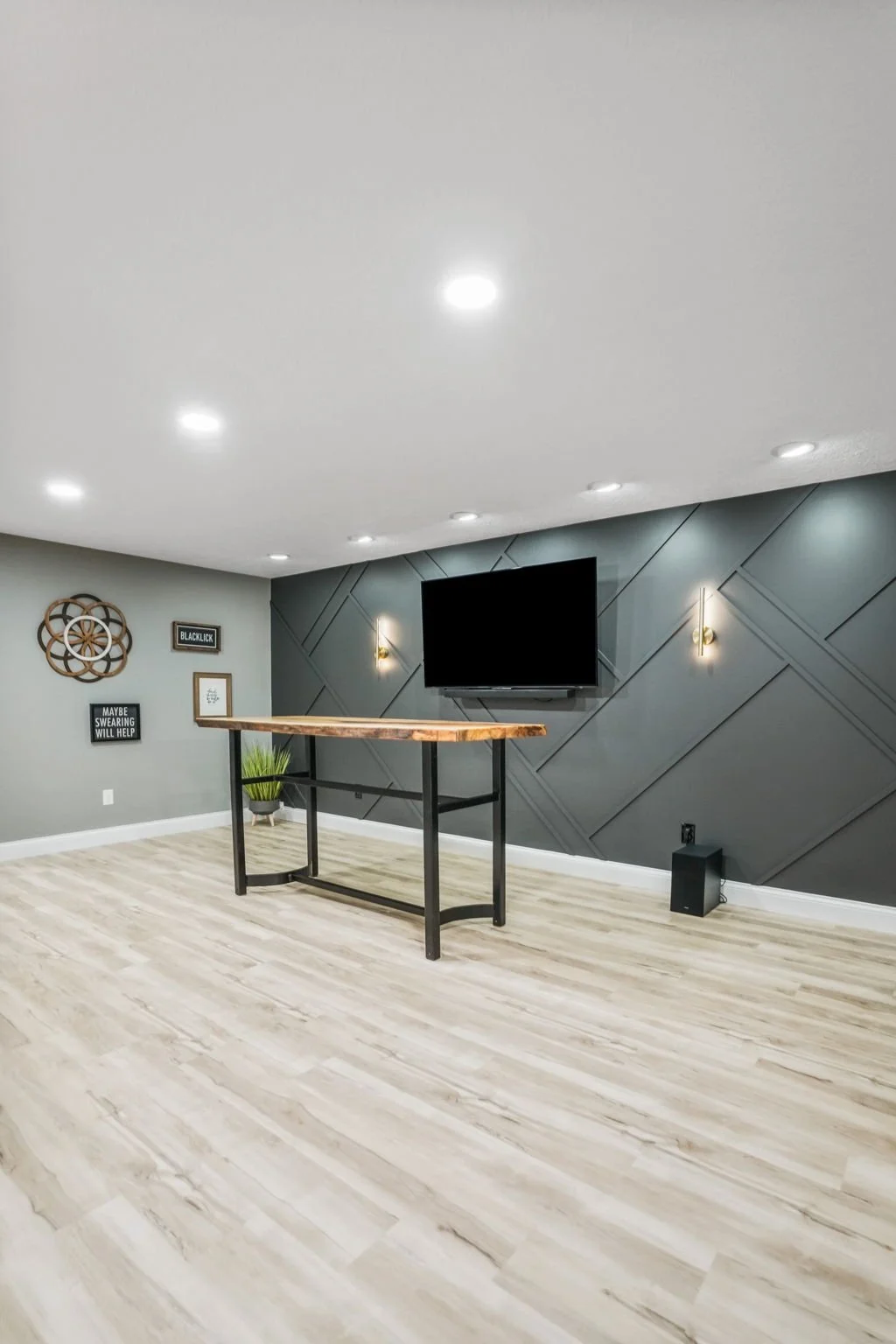 Modern living room with a mounted flat-screen TV, gray textured accent wall, light wood flooring, small black speaker, and wall decor including a wooden geometric wall art, framed picture, and wall sign.