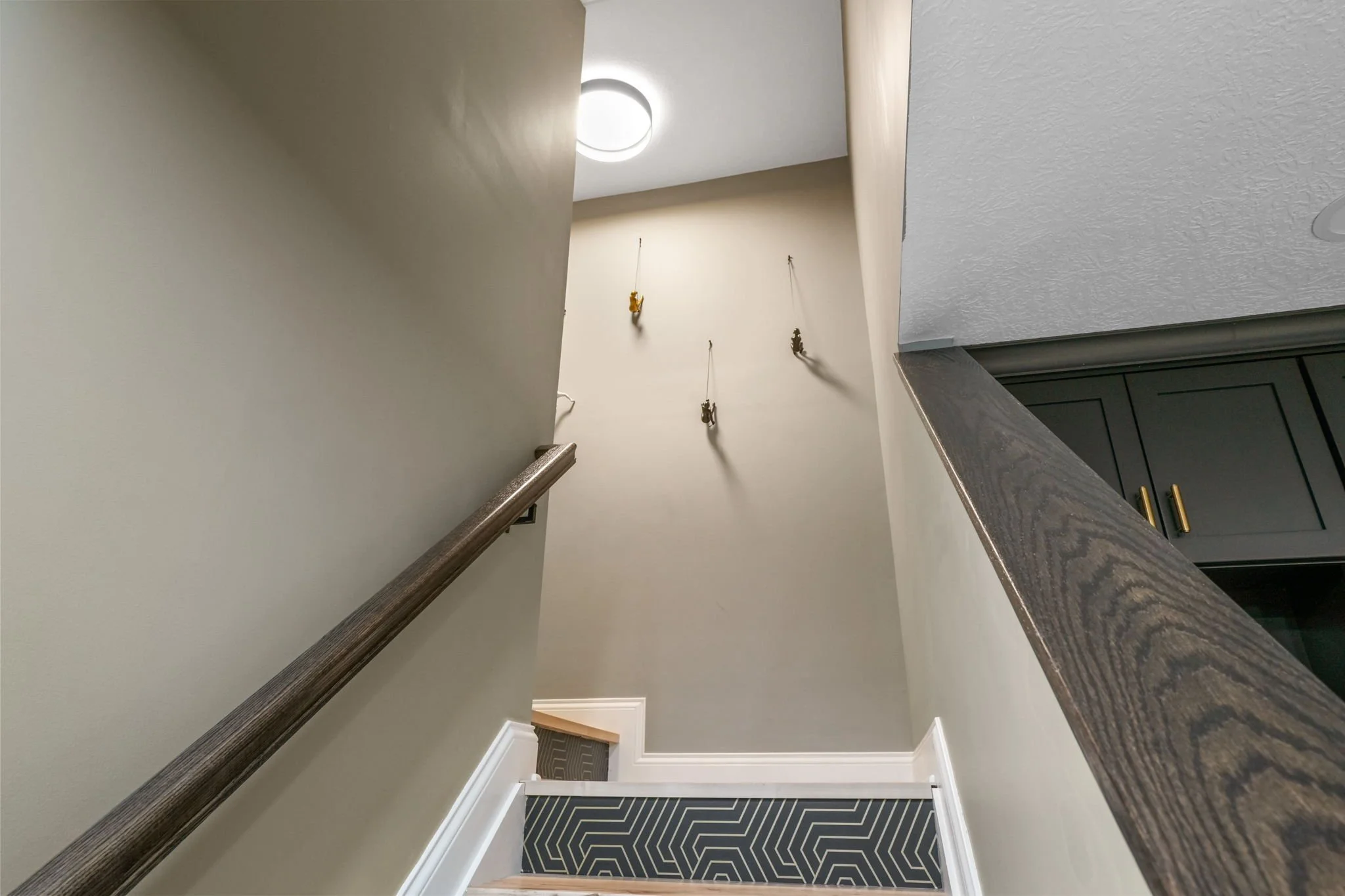 View of a staircase viewed from the top, with a beige wall on the left and a black textured narrow railing on the right, facing down towards a small landing with patterned black and white carpet, and a beige wall with three hooks and hanging keychain