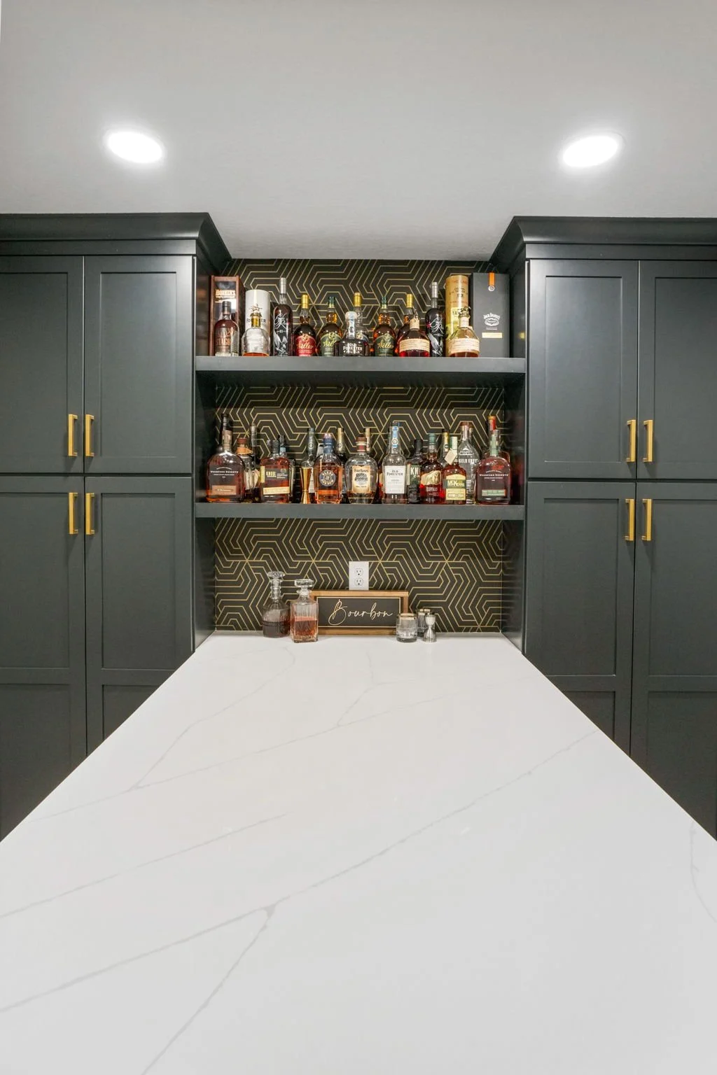 Home bar with dark cabinets, gold handles, black patterned backsplash, and various liquor bottles on open shelves.