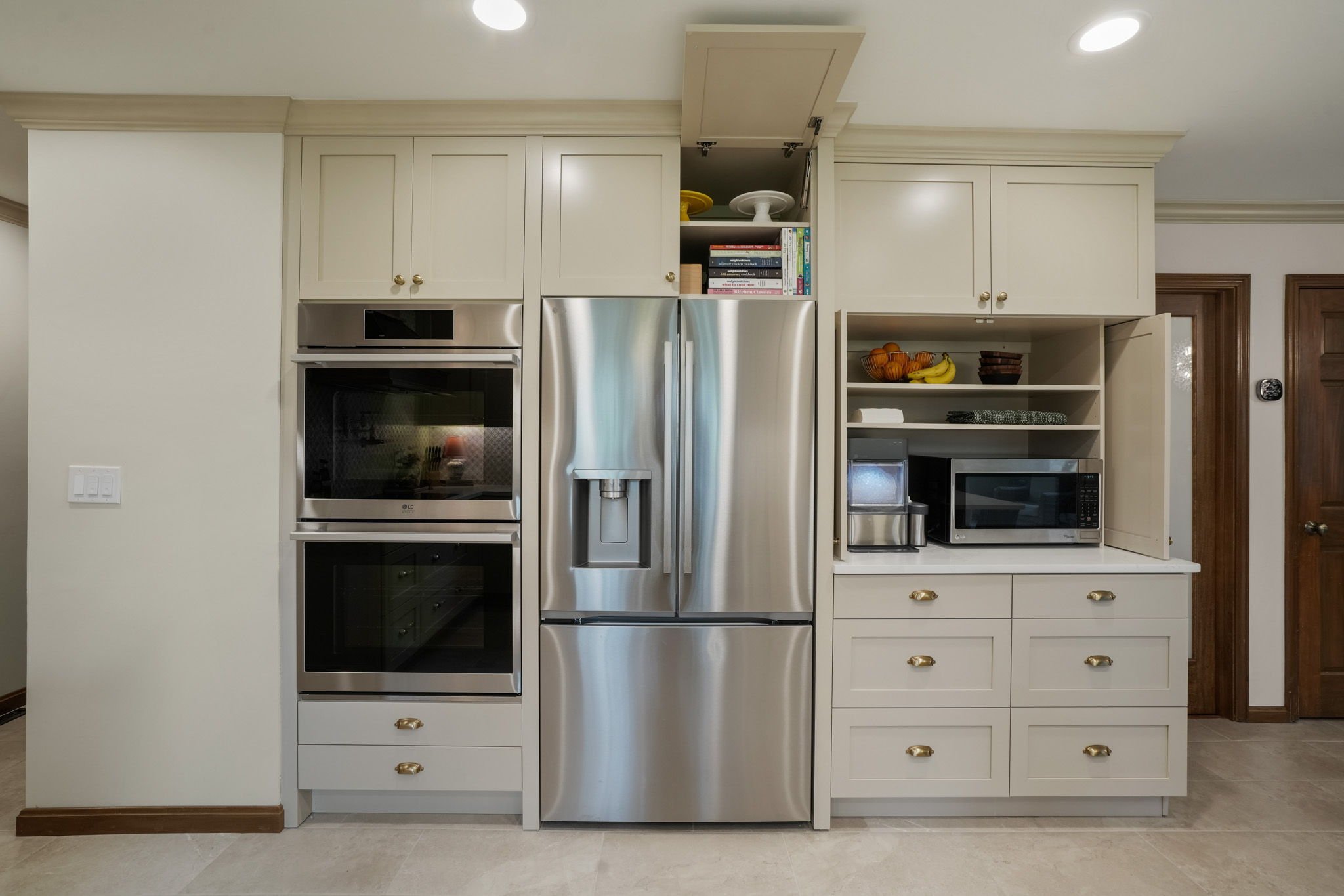 Kitchen with stainless steel refrigerator, double oven, microwave, open cabinets displaying fruit and books, and beige cabinetry with gold handles.