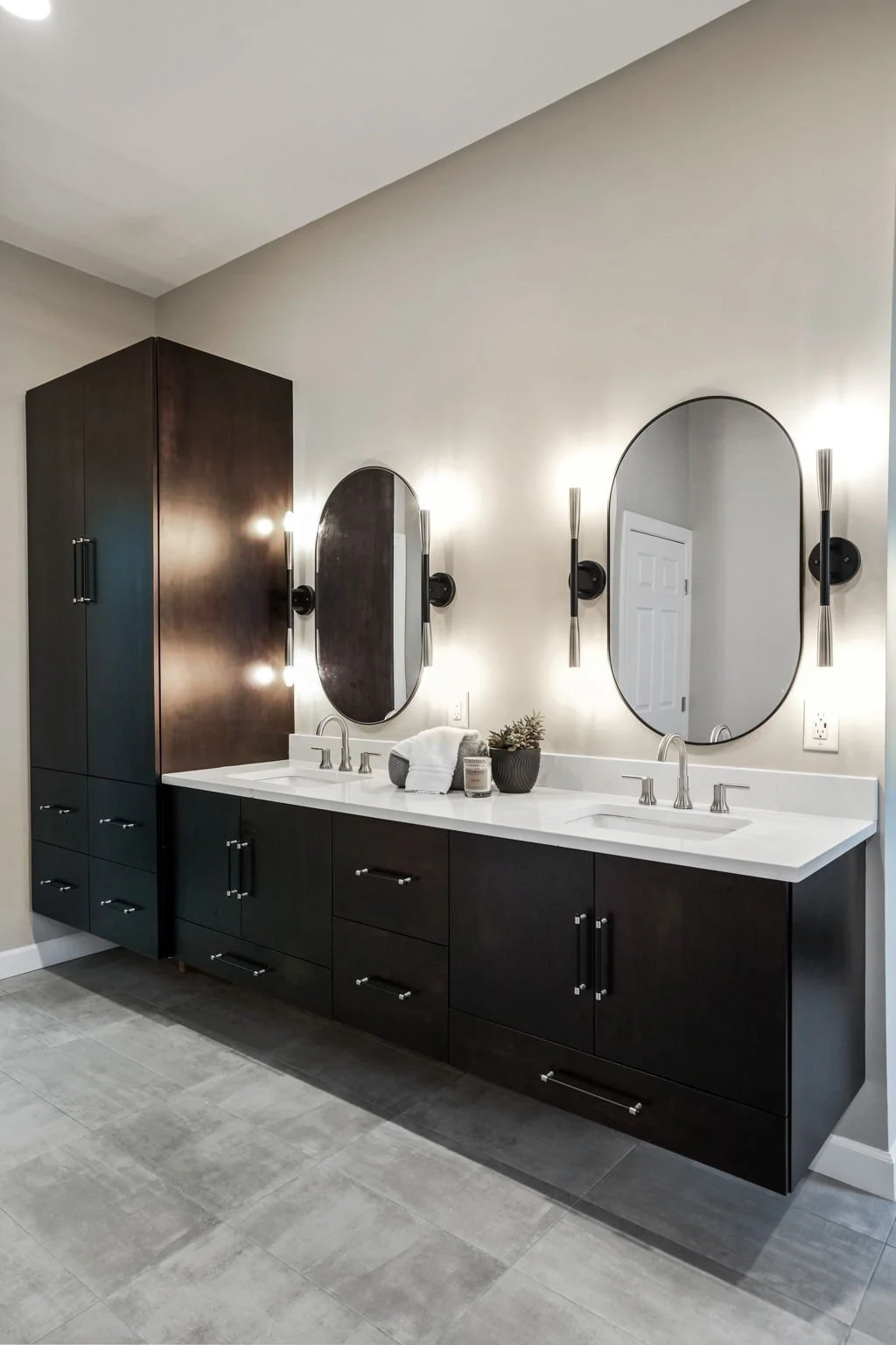 Modern bathroom vanity with two oval mirrors, black cabinets, white countertops, and dual sinks, illuminated by wall-mounted light fixtures.