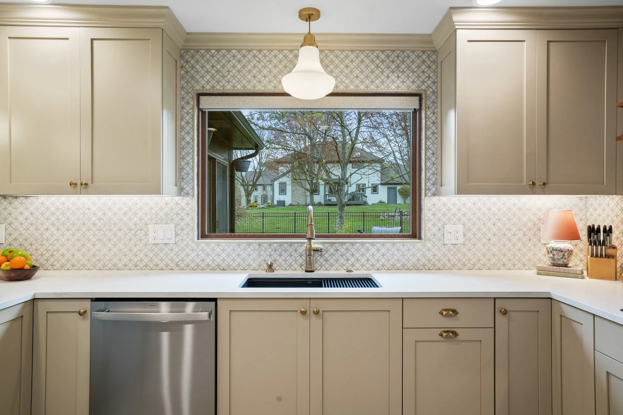 Kitchen with beige cabinets, a window showing a backyard with trees and houses, and a countertop with a bowl of fruit, a small lamp, and a knife block.