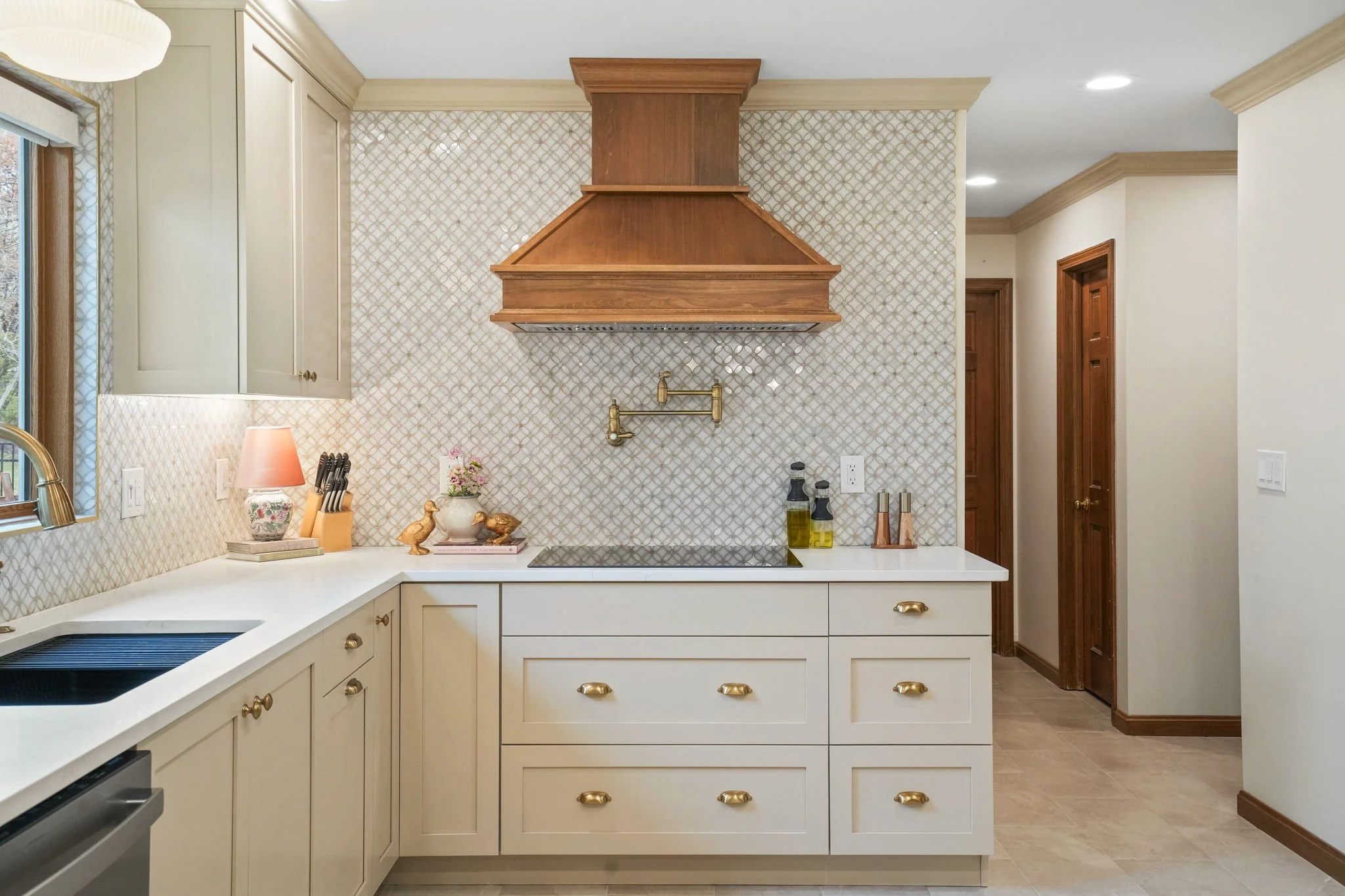Kitchen with white cabinets, gold hardware, and a patterned backsplash. A wooden range hood above the cooktop, with a brass pot filler, and decorative items including a pink lamp, books, and duck figurines on the countertop. There are windows with wood trim and recessed lighting.