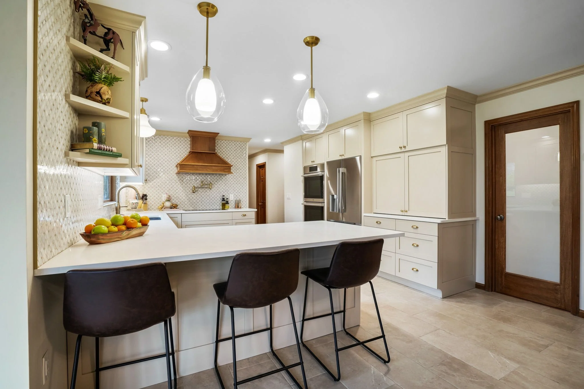 Modern kitchen with white cabinets, a breakfast bar with three dark chairs, pendant lights, stainless steel refrigerator, and a wooden door.