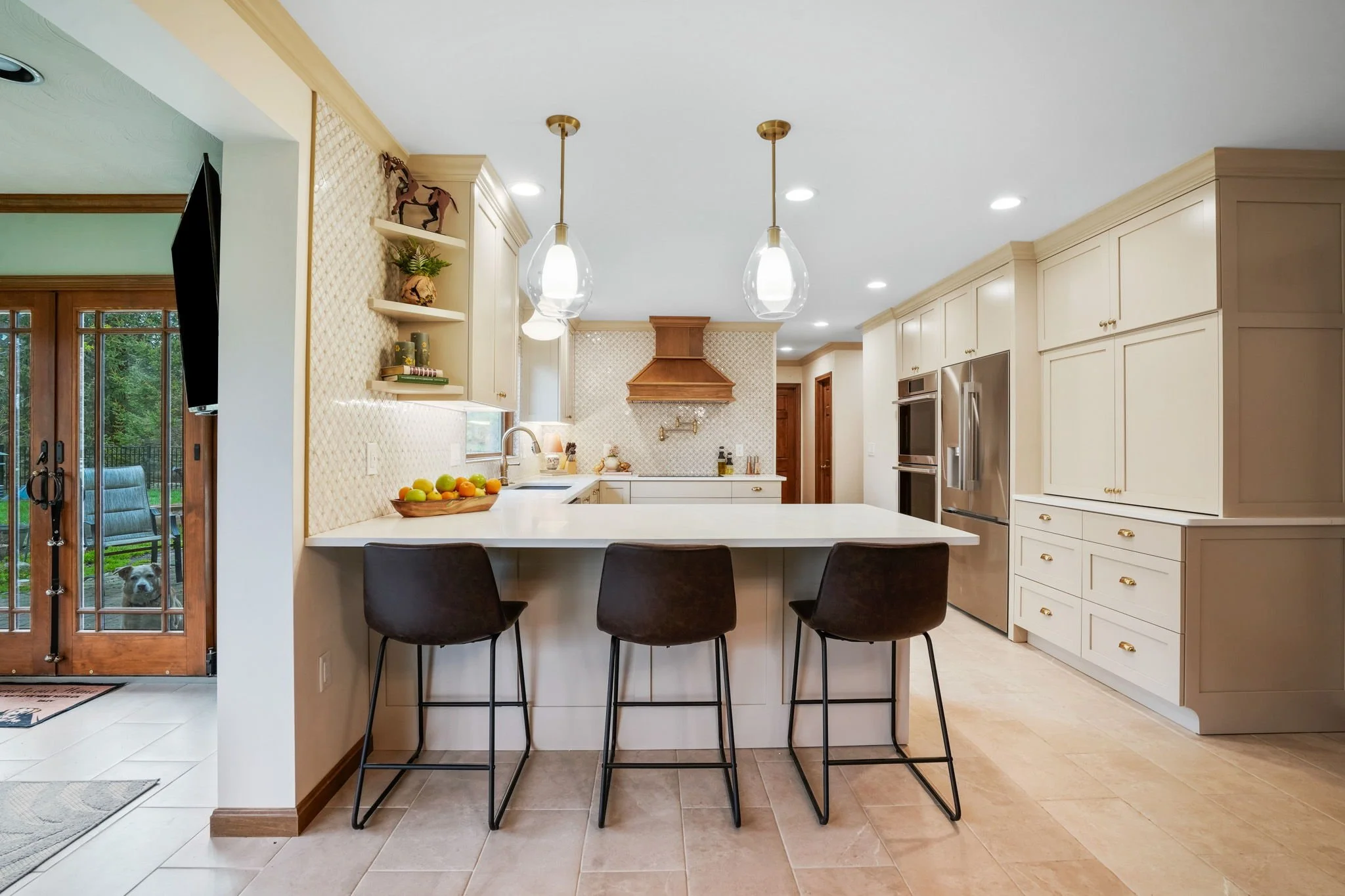Modern kitchen with white cabinets, stainless steel refrigerator, and island with three black chairs. Recessed lighting and pendant lights hang above the island. Sliding glass door leads outside, where a dog can be seen.