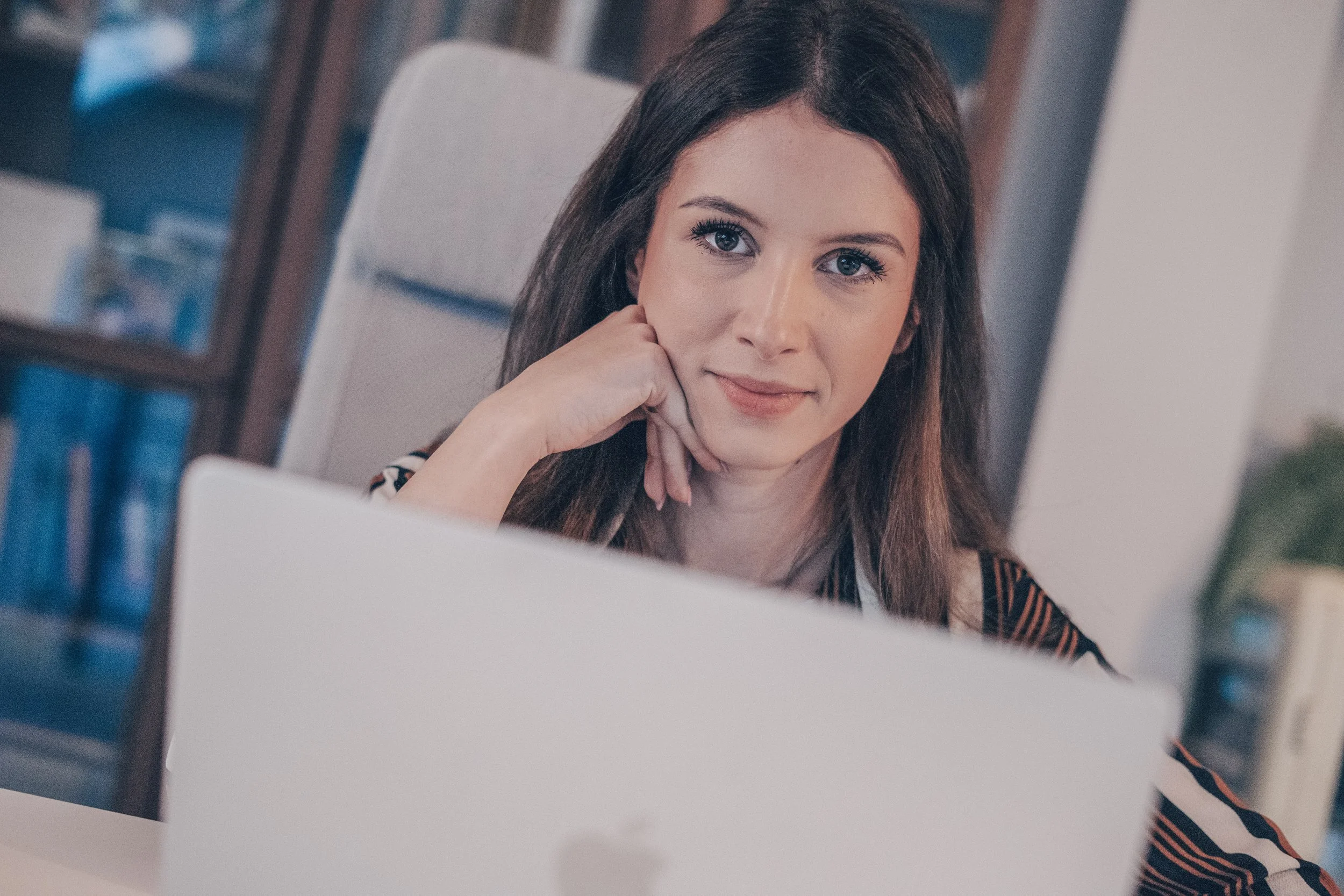 A woman with long dark hair and blue eyes sitting at a desk with a laptop, resting her chin on her hand, looking at the camera, in an office or home setting.