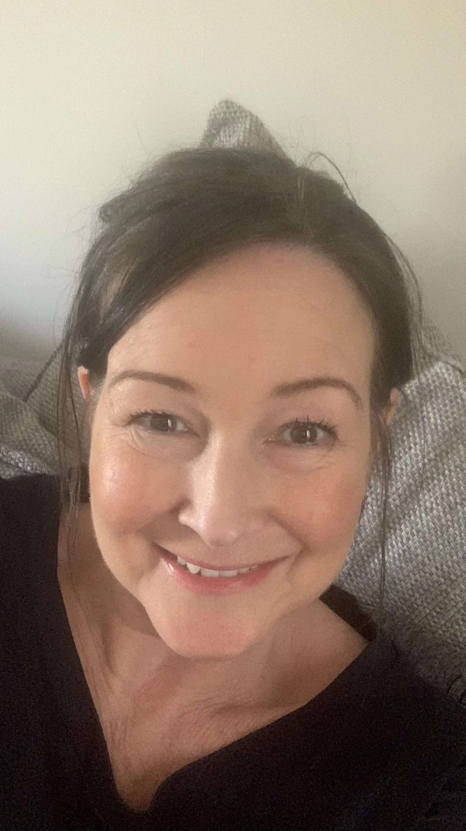 A smiling woman with dark hair, wearing a black top, sitting on a gray textured couch.