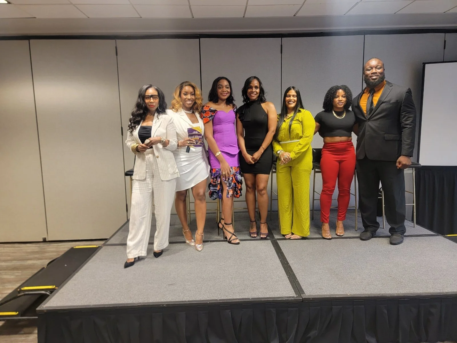 Group of eight diverse professionals, seven women and one man, standing on a stage dressed in business attire at a conference or event.