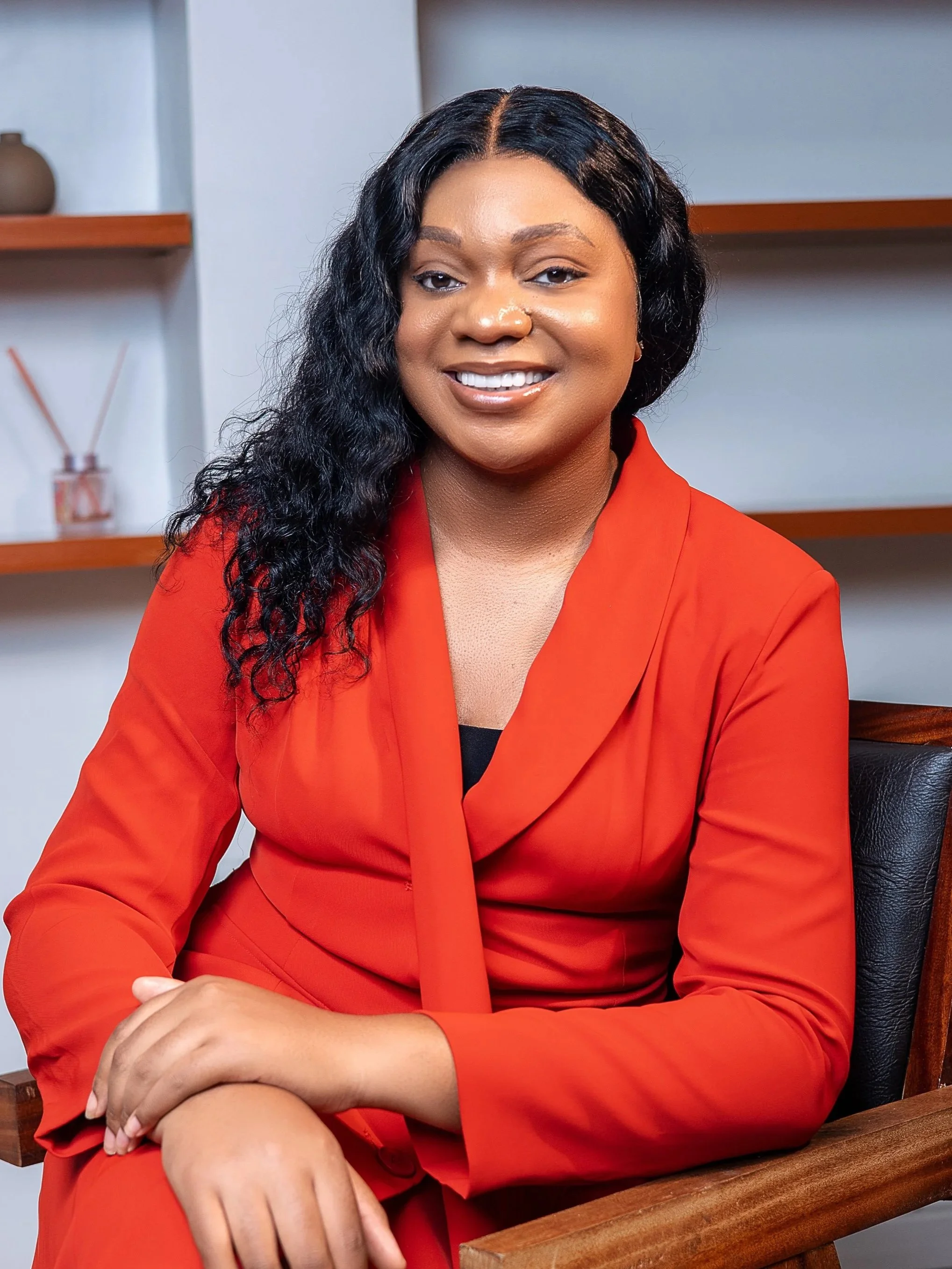 image of Unwanaobong Udoko Therapist, A woman with long curly black hair sitting in a wooden chair, wearing a red blazer, smiling warmly in a modern, minimalistic room.