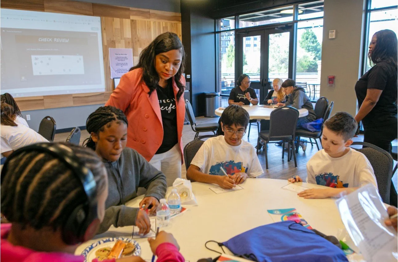 Children sitting at a round table in a classroom or community center, engaging in an activity, with a woman standing nearby supervising. In the background, there are other groups of people, large windows, and a projector screen on a wooden wall.