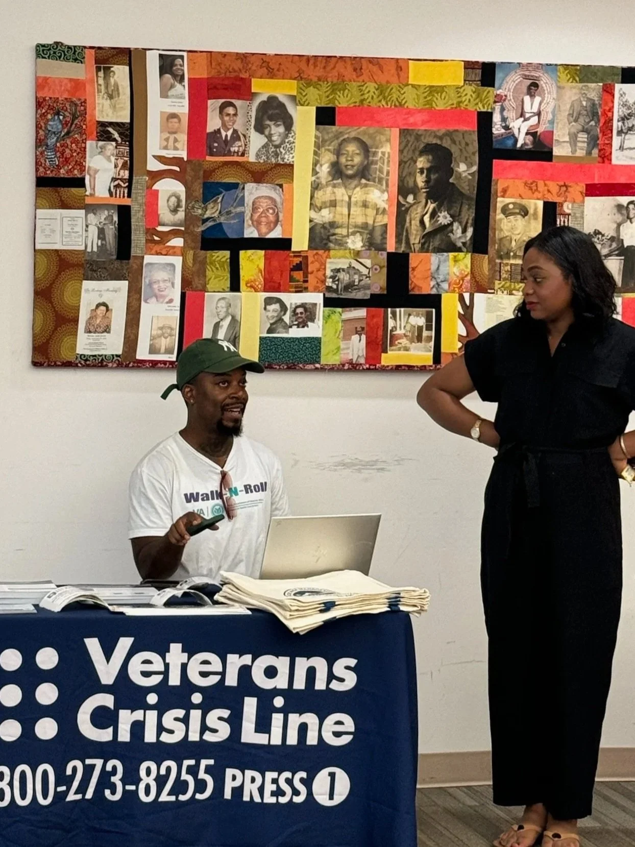 A man and a woman conversing next to a table with a Veterans Crisis Line banner in a room with a colorful photo collage on the wall.