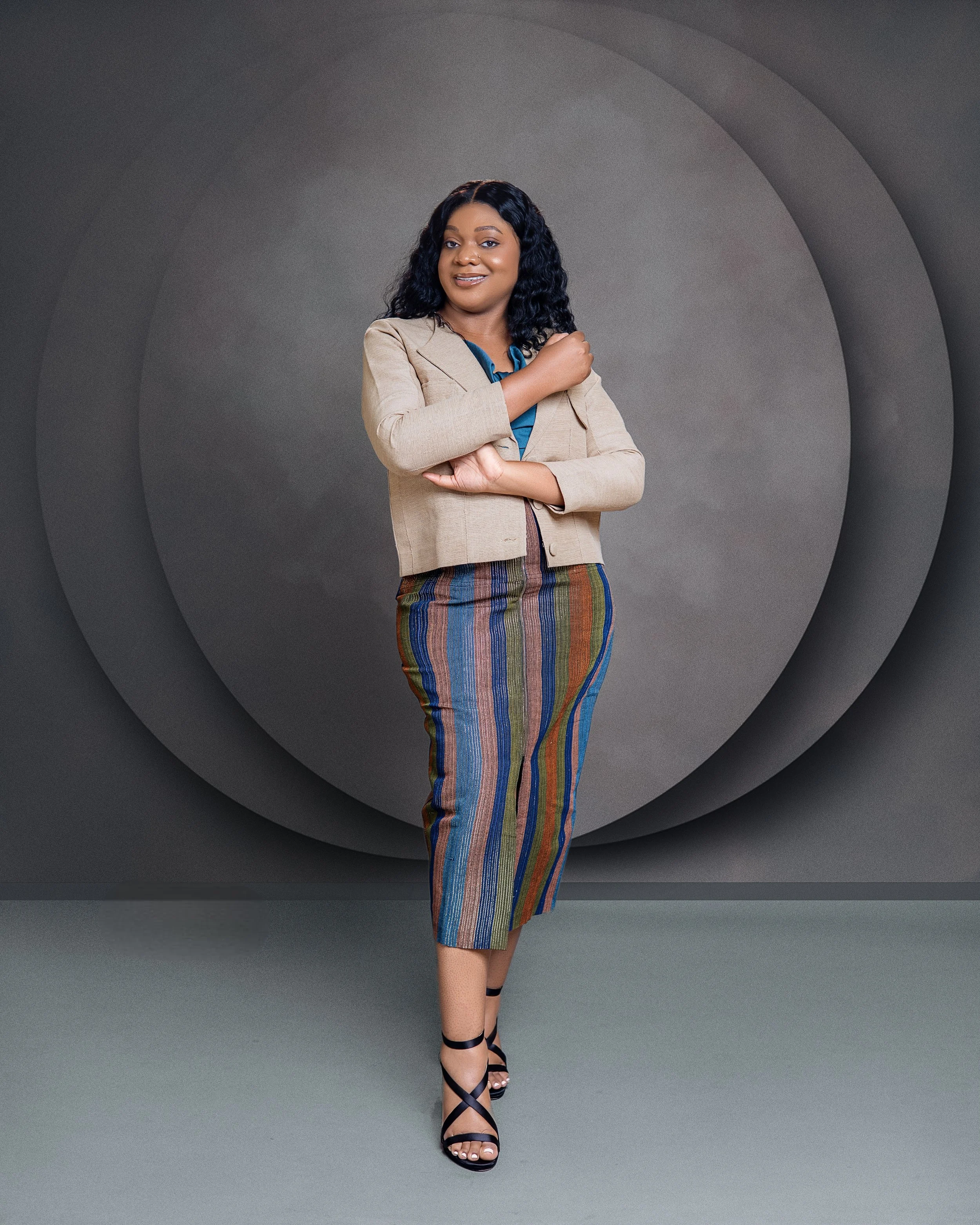 A woman with curly black hair smiling confidently, wearing a beige blazer, a blue blouse, colorful striped pants, and black strappy heels, standing with arms crossed in front of a gray, layered circular background.
