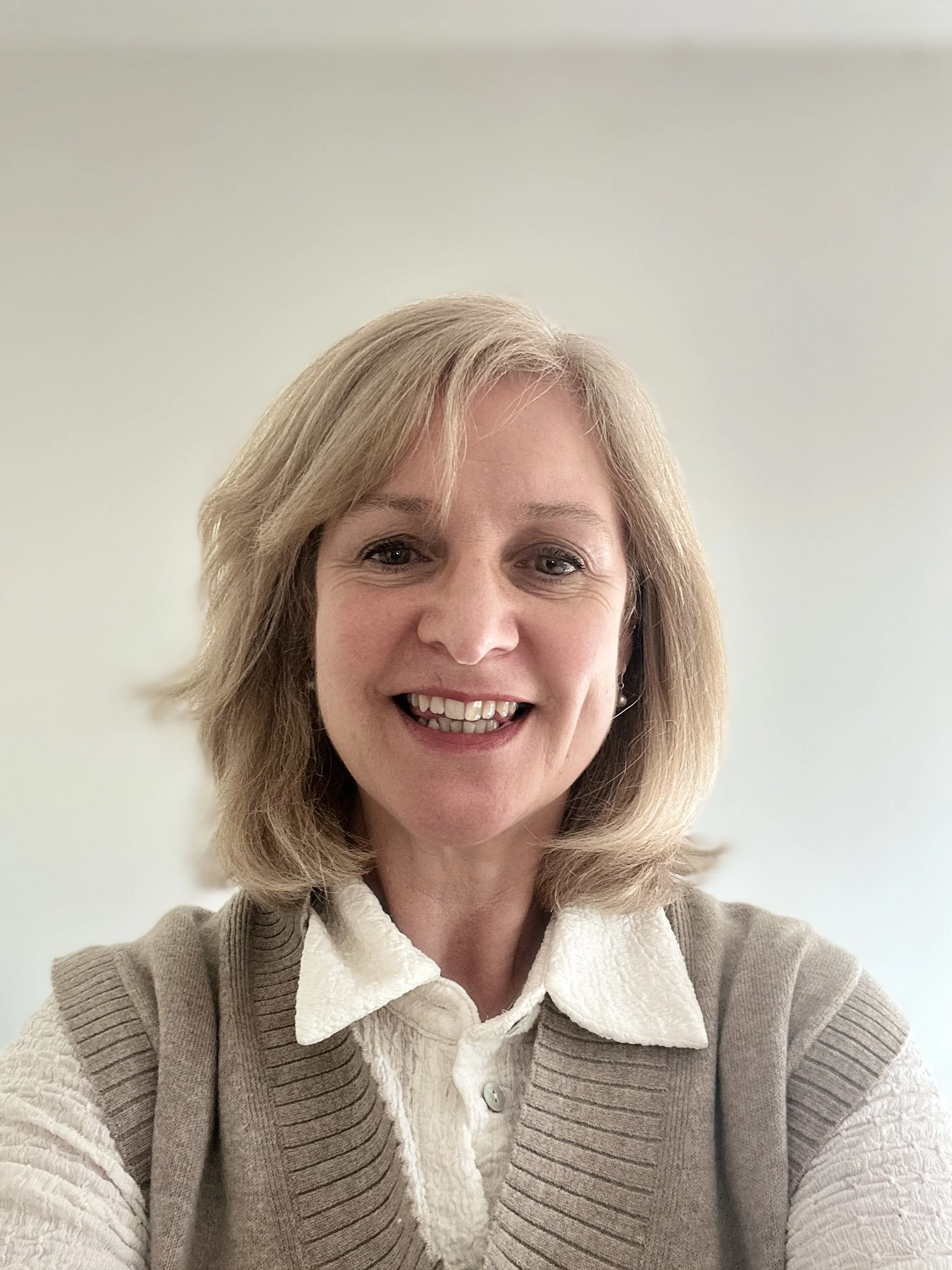 A woman with blonde hair smiling, wearing a cream-colored shirt and a brown vest, against a plain light-colored background.