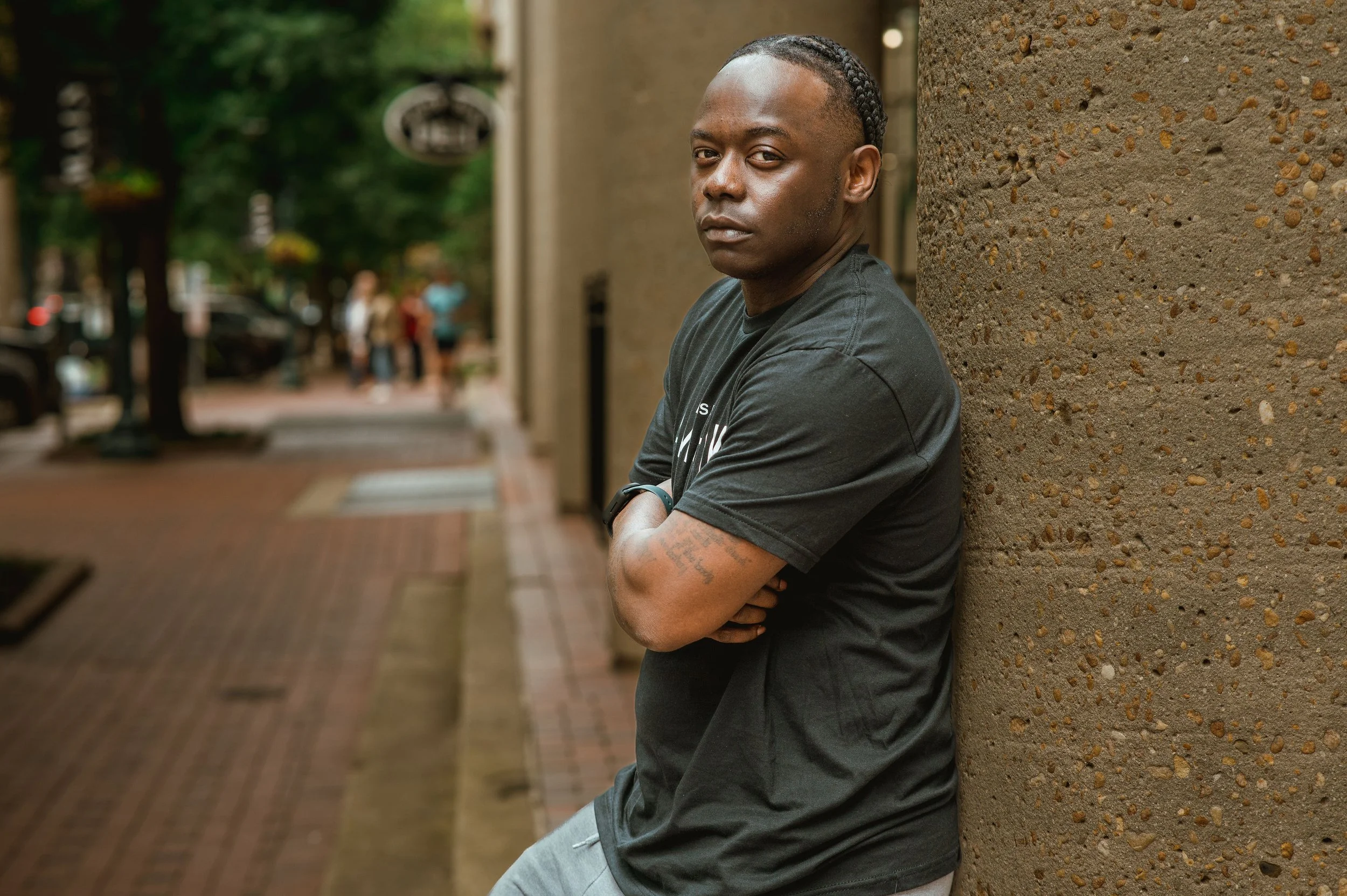 Young man with braided hair and tattoos leaning against a textured wall on a city sidewalk, crossing his arms and looking into the camera.