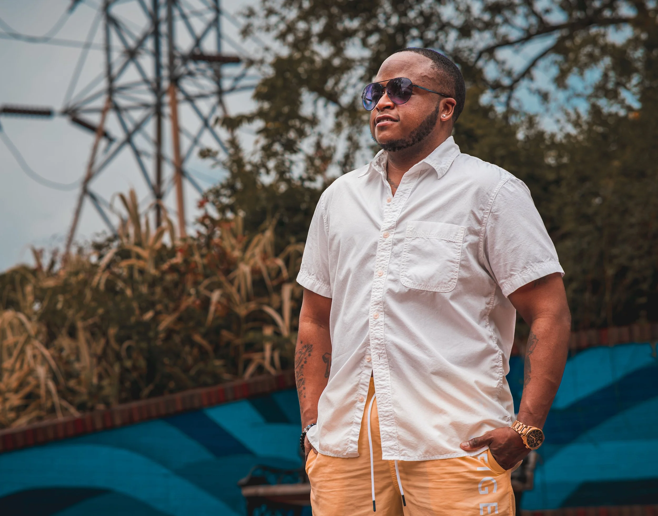 A young man wearing dark sunglasses, a white short-sleeved button-up shirt, beige shorts, and a gold watch, standing outdoors with his hands in his pockets. There is greenery and a blue structure in the background.