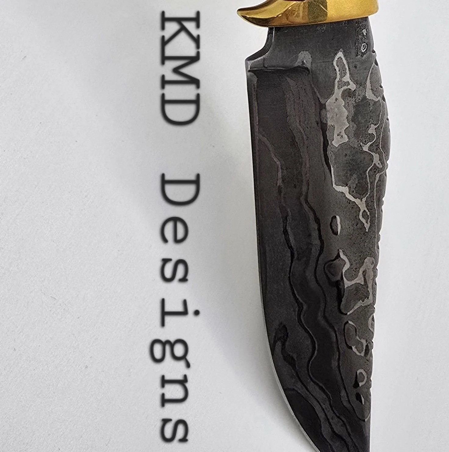 A black and gray patterned knife with a gold-colored handle resting on a white surface. The words "Sunday, De S. T. VIII" are printed vertically on the left side of the image.