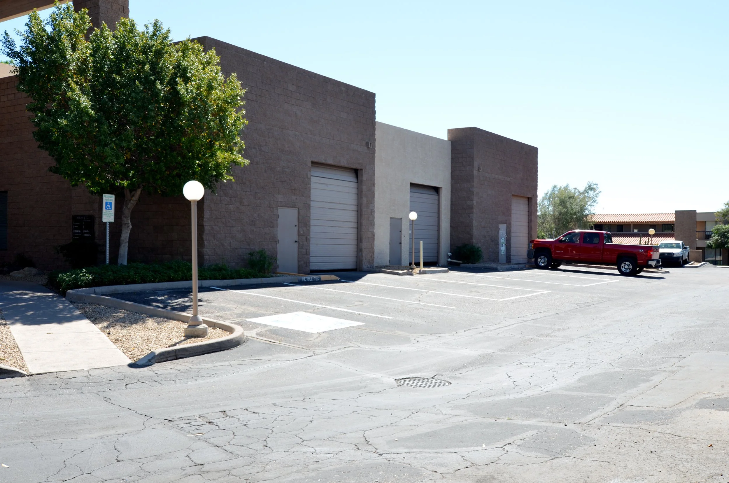 Empty parking lot outside a beige and brown building with trees, street lamps, and parked cars.