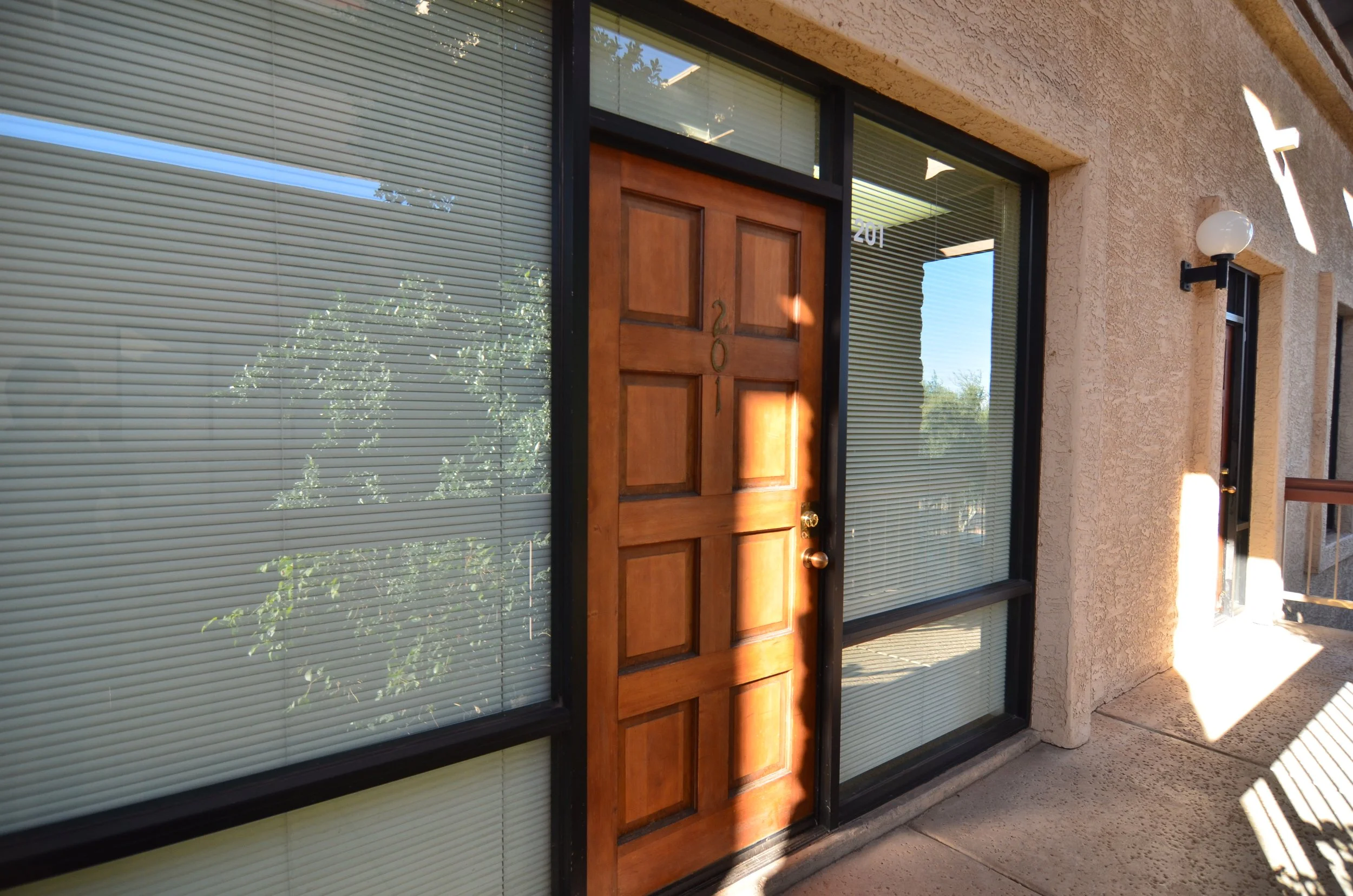 Front view of a house with a wooden door and large windows with closed blinds, exterior wall textured and beige, with outdoor wall mounted light fixtures, and sunlight casting shadows.