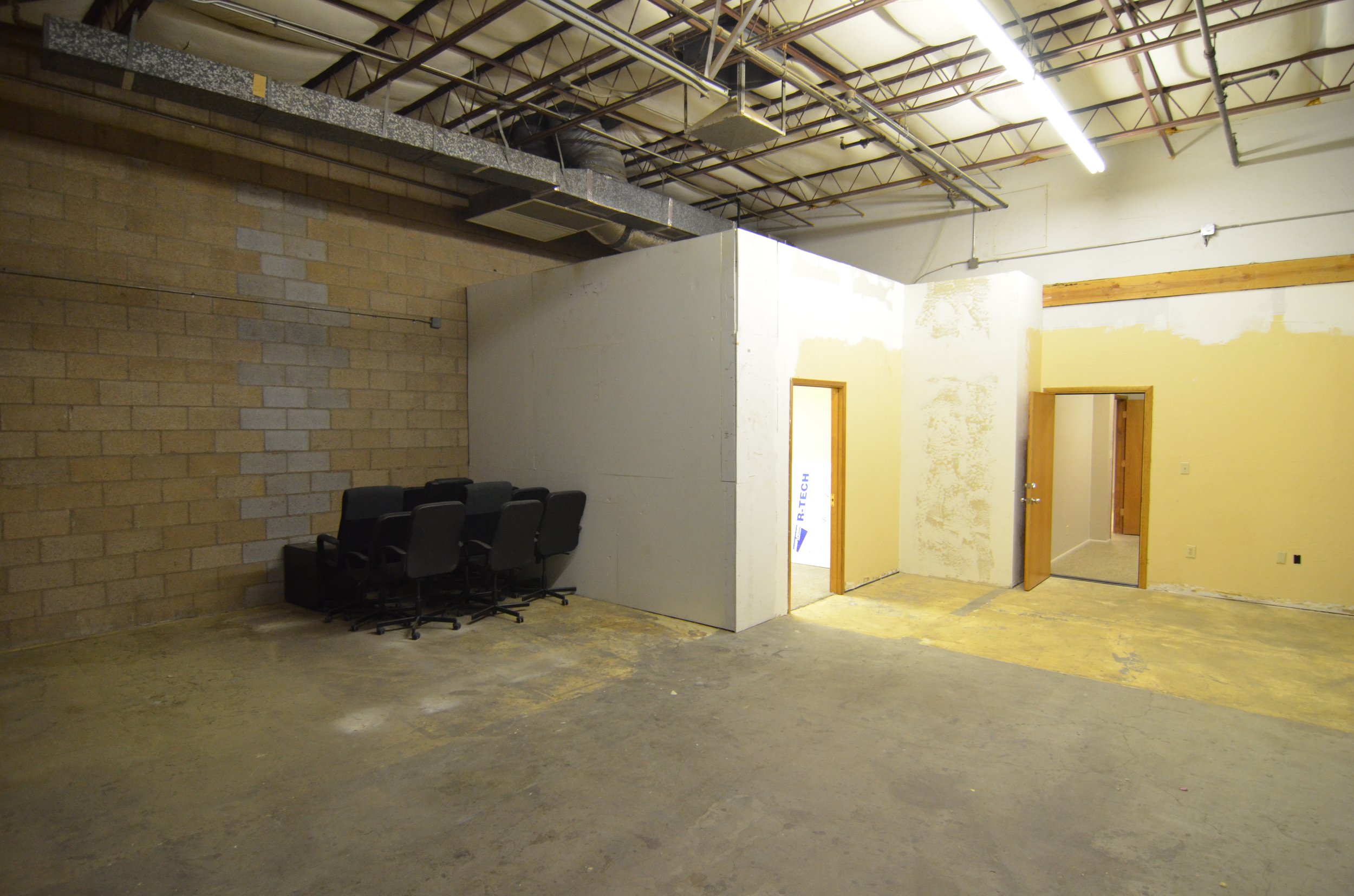 Empty room with concrete floor, brick wall on the left, yellow walls, and exposed ceiling with pipes and beams. There are some black chairs arranged in a row, and a white partition. Two doorways are visible, one with a door and one open without a doo