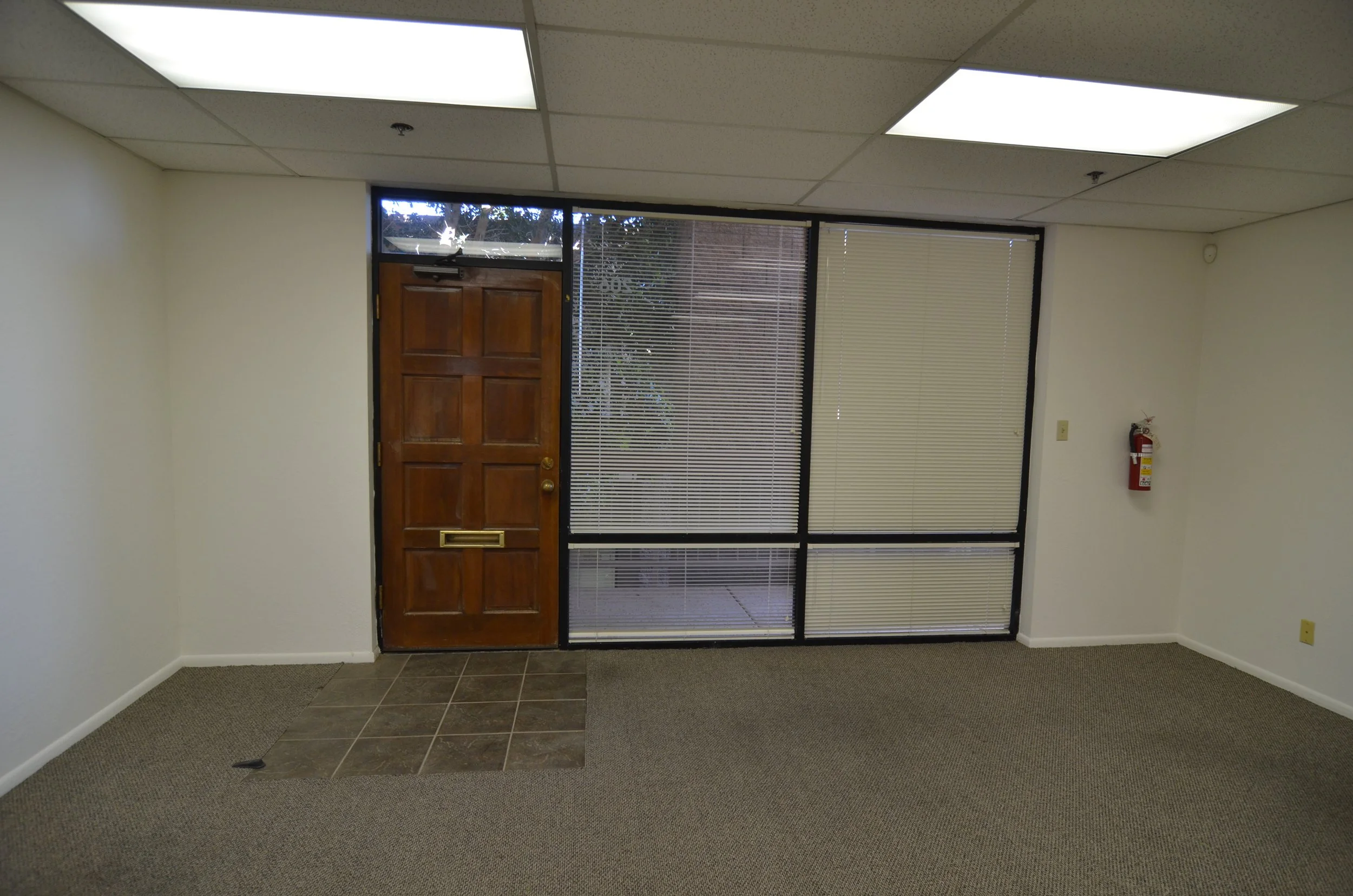 Empty office space with a wooden door, large windows with blinds, ceiling fluorescent lights, a fire extinguisher on the wall, and a mix of carpet and tile flooring.