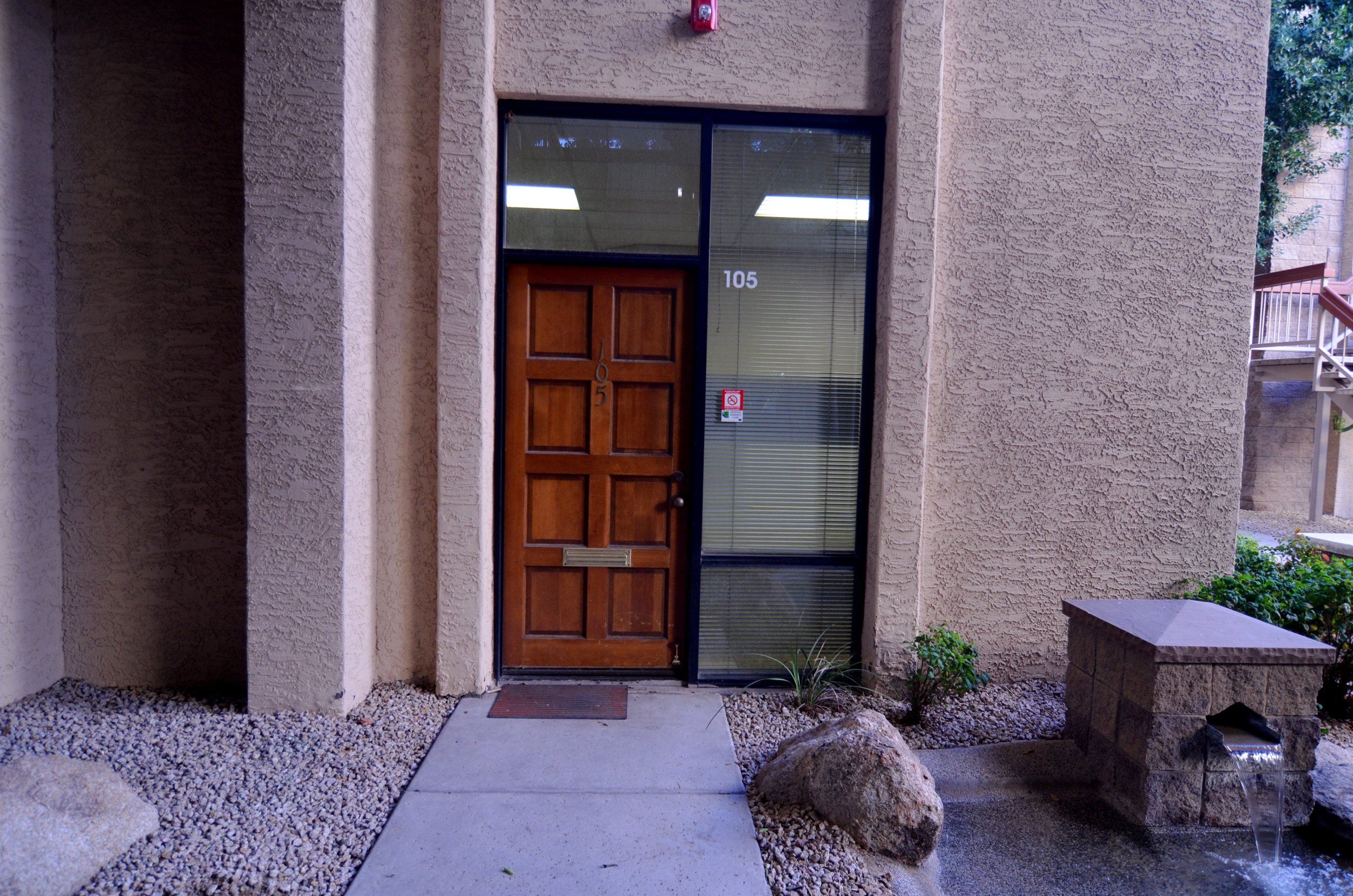 An apartment building entrance with a wooden door labeled 105, a glass window, and a small red fire alarm. There are rocks, gravel, and a small garden with a plant outside the entrance.