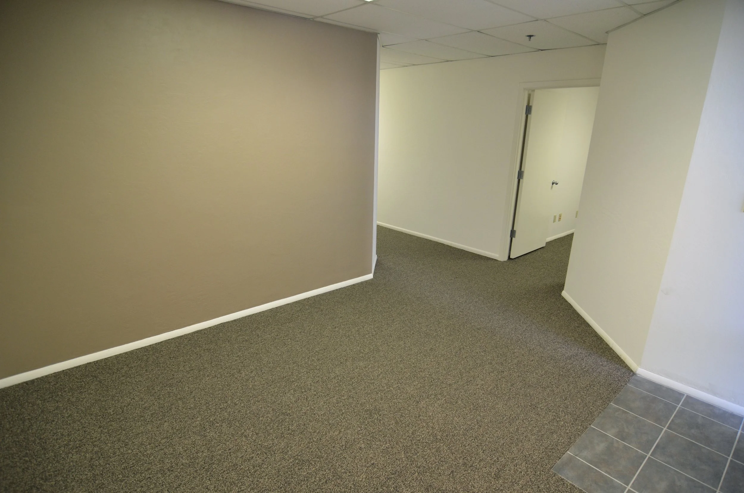 An interior hallway with beige walls, gray carpeted floor, and a drop ceiling. There is an open door on the right side leading to another room with a floor tile section visible, and the hallway has a simple, clean appearance.