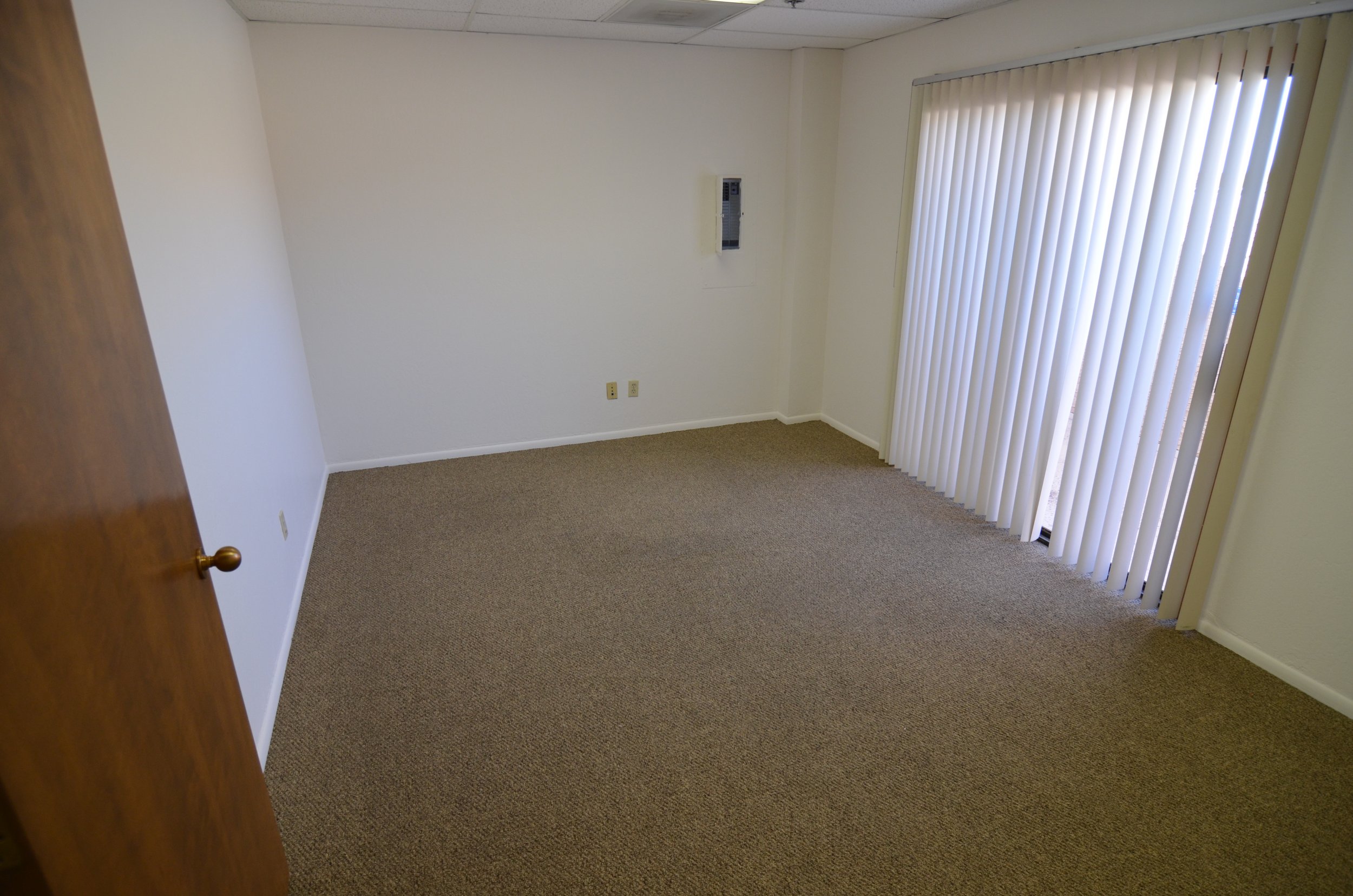 Empty room with beige carpet, white walls, vertical blinds covering a window, and an air conditioning unit on the wall.