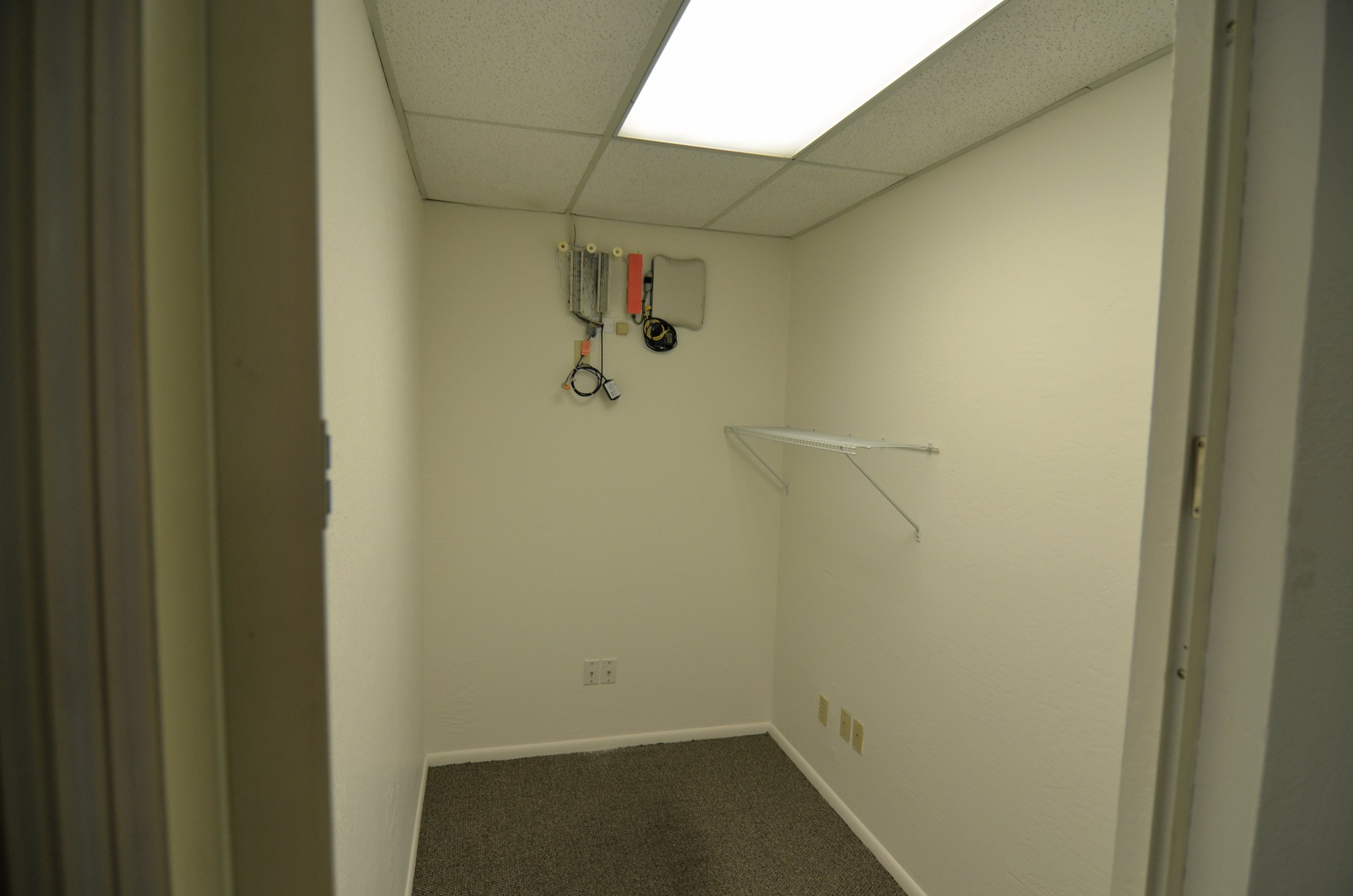 Empty beige-walled office or storage room with a ceiling-mounted fluorescent light, a white wire shelf on the right wall, and electrical outlets at the bottom.