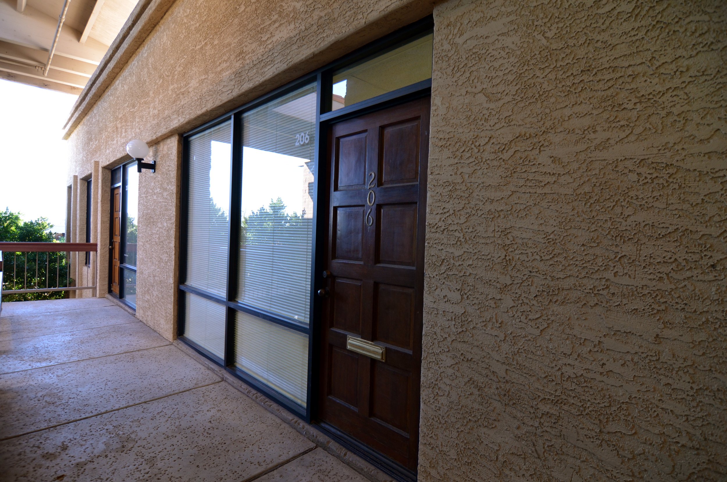 Officespace entrance with brown door, large windows with blinds, off-white textured exterior wall, and a wall-mounted light fixture.