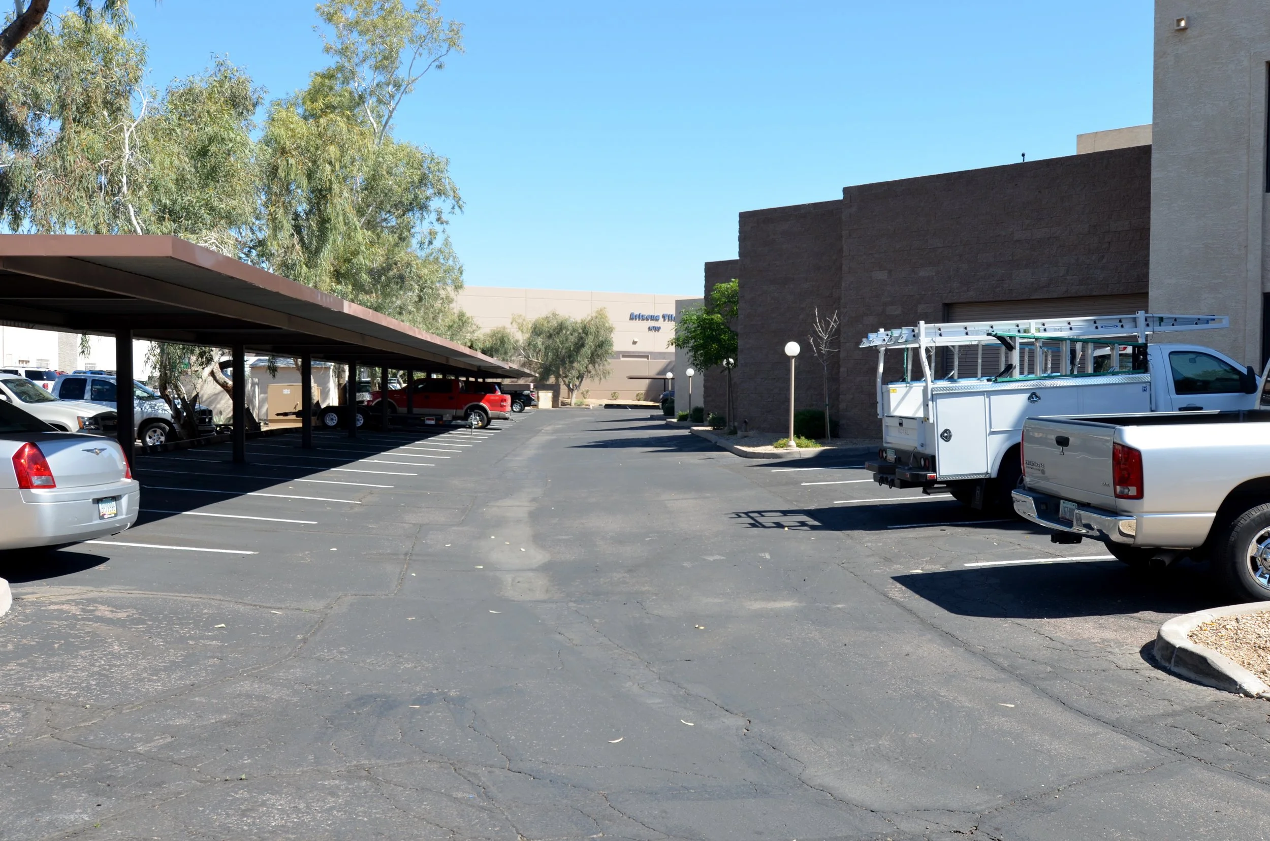 Empty parking lot with cars parked under a covered structure, trees, and a building in the background.